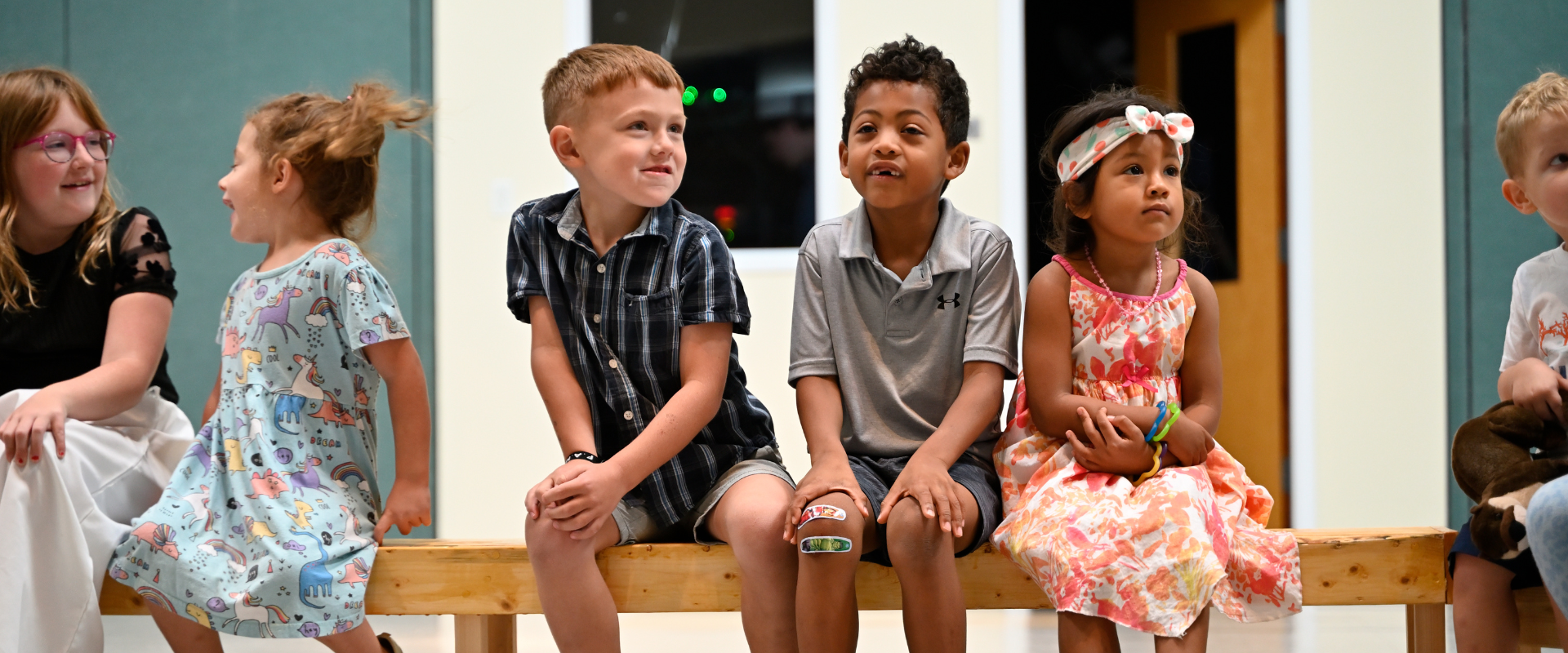 Group of young children sitting on a wooden bench during a performance or event, in a brightly lit room.