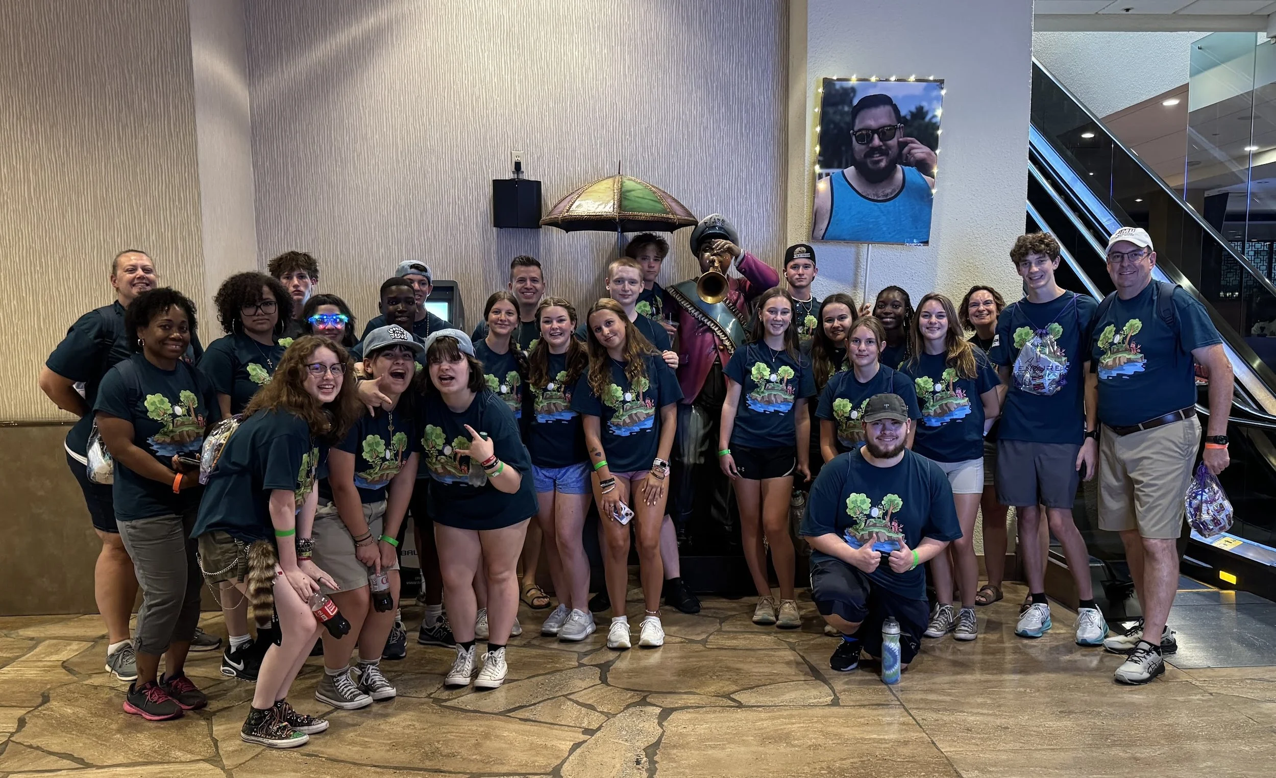 A large group of young people and a few adults posing for a photo indoors. They are smiling and standing together. Some are holding souvenirs, and one person is playing a trumpet. There is a large framed photograph of a man wearing sunglasses on the 