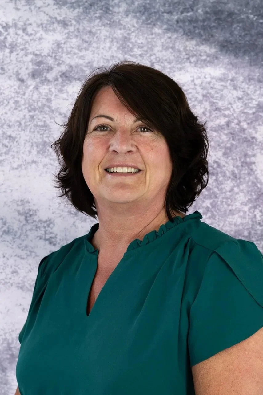 A woman with short dark brown hair and a teal blouse standing in front of a gray textured background.