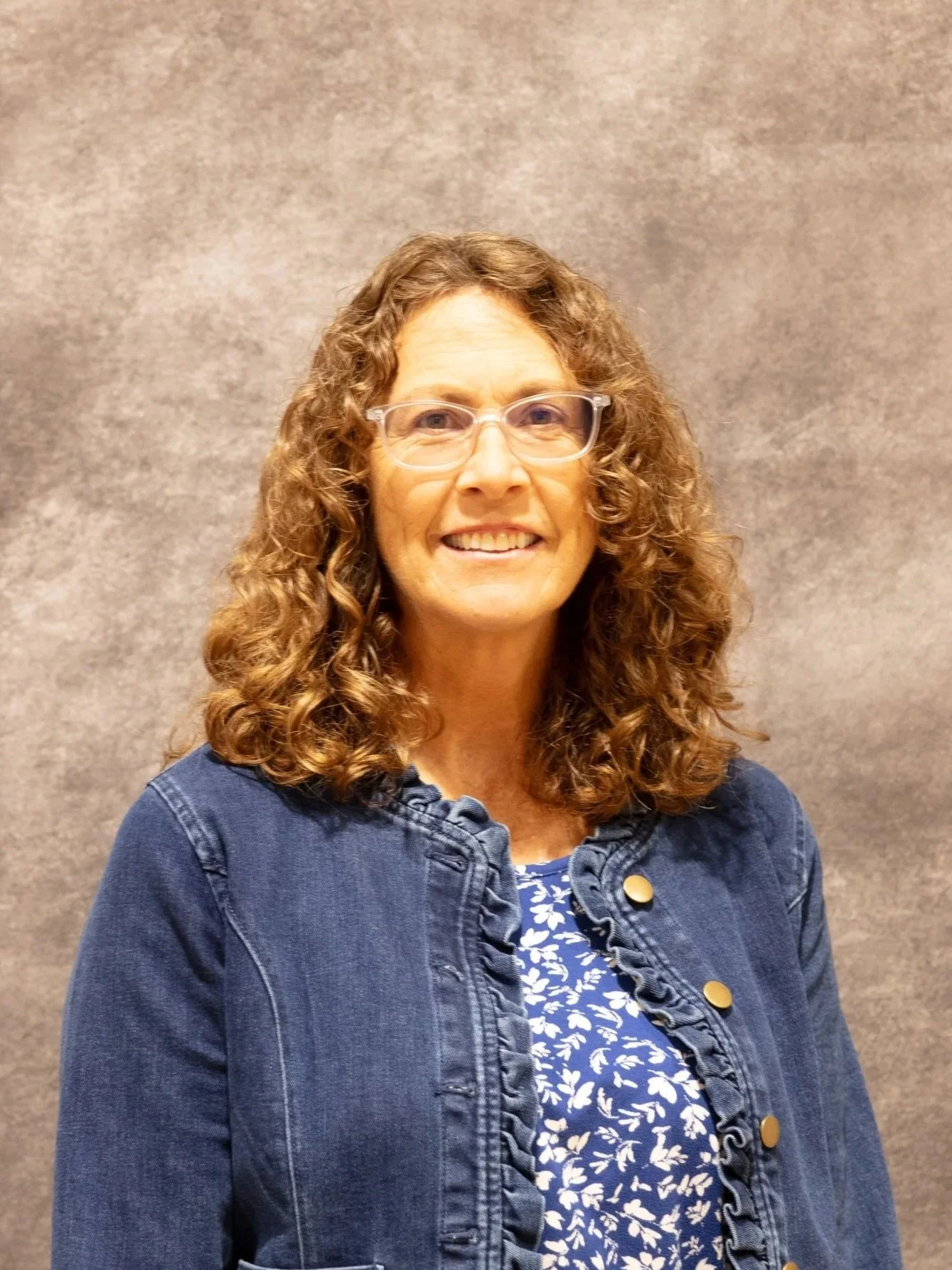 A woman with glasses and curly hair smiling, wearing a denim jacket over a blue and white patterned blouse, standing against a gray textured background.