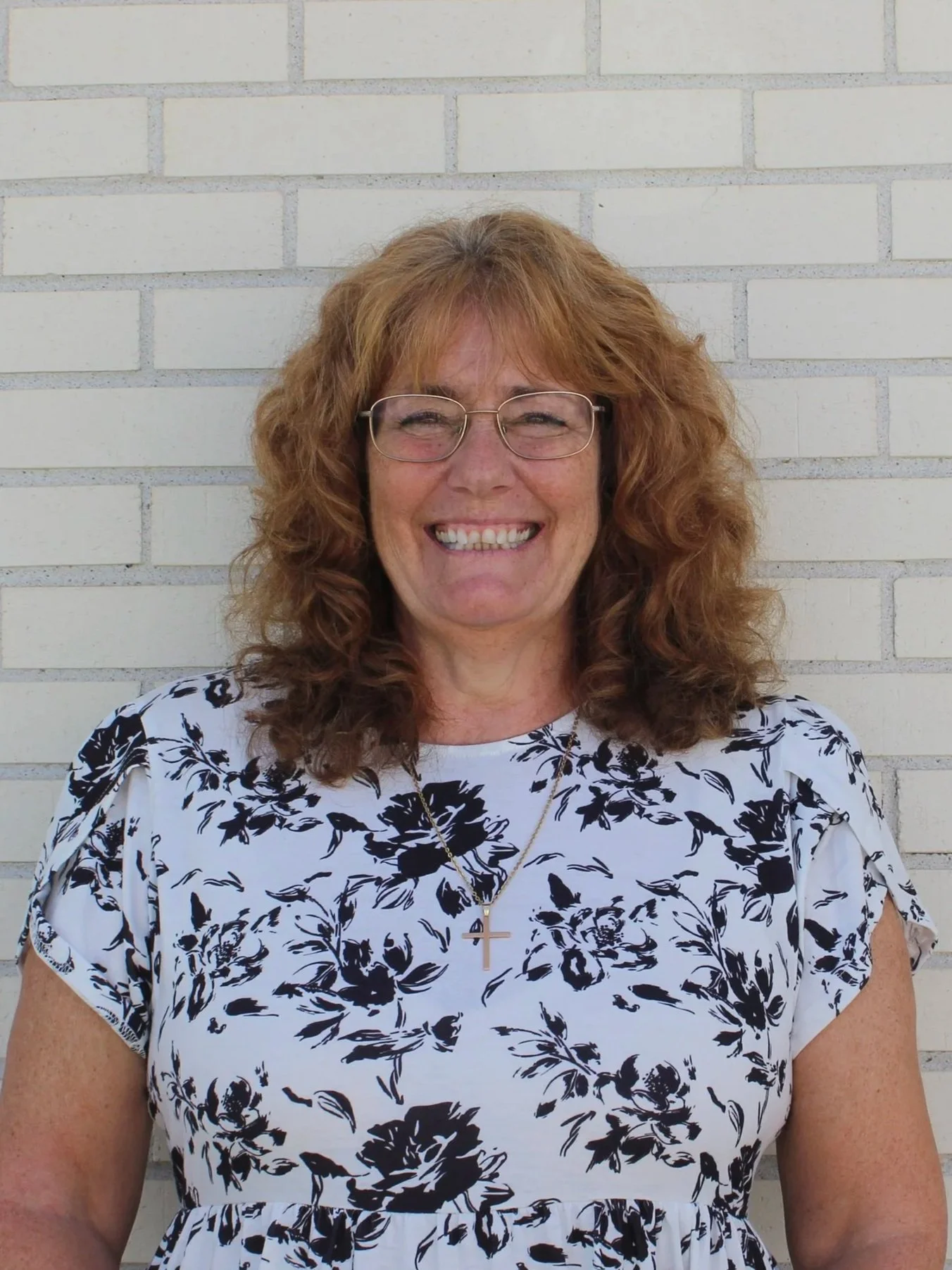 A woman with curly red hair and glasses smiling, wearing a white dress with black floral patterns, standing in front of a white brick wall.