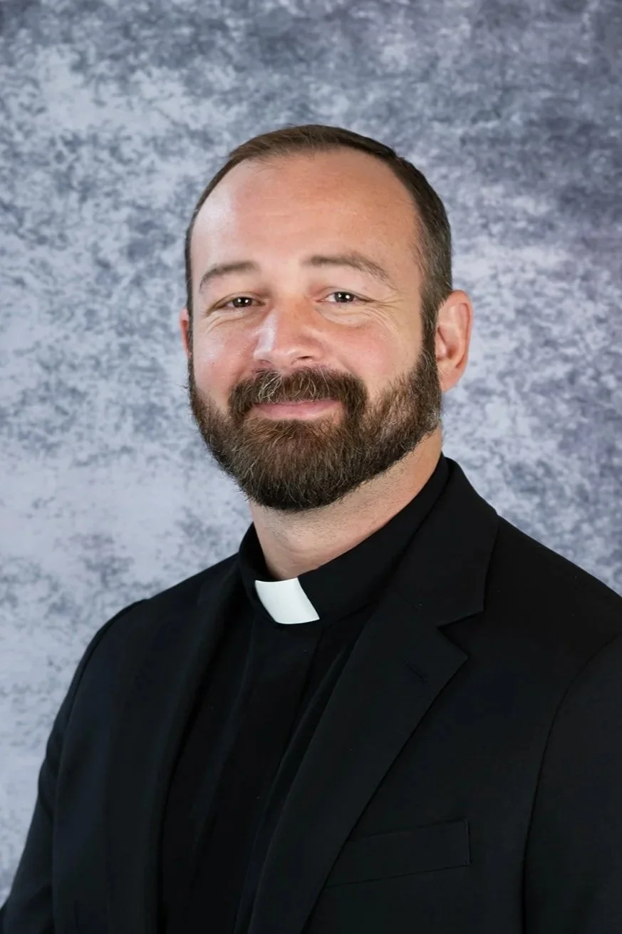 Portrait of a bearded man wearing a priest's collar, dressed in a black suit, standing against a textured gray background.