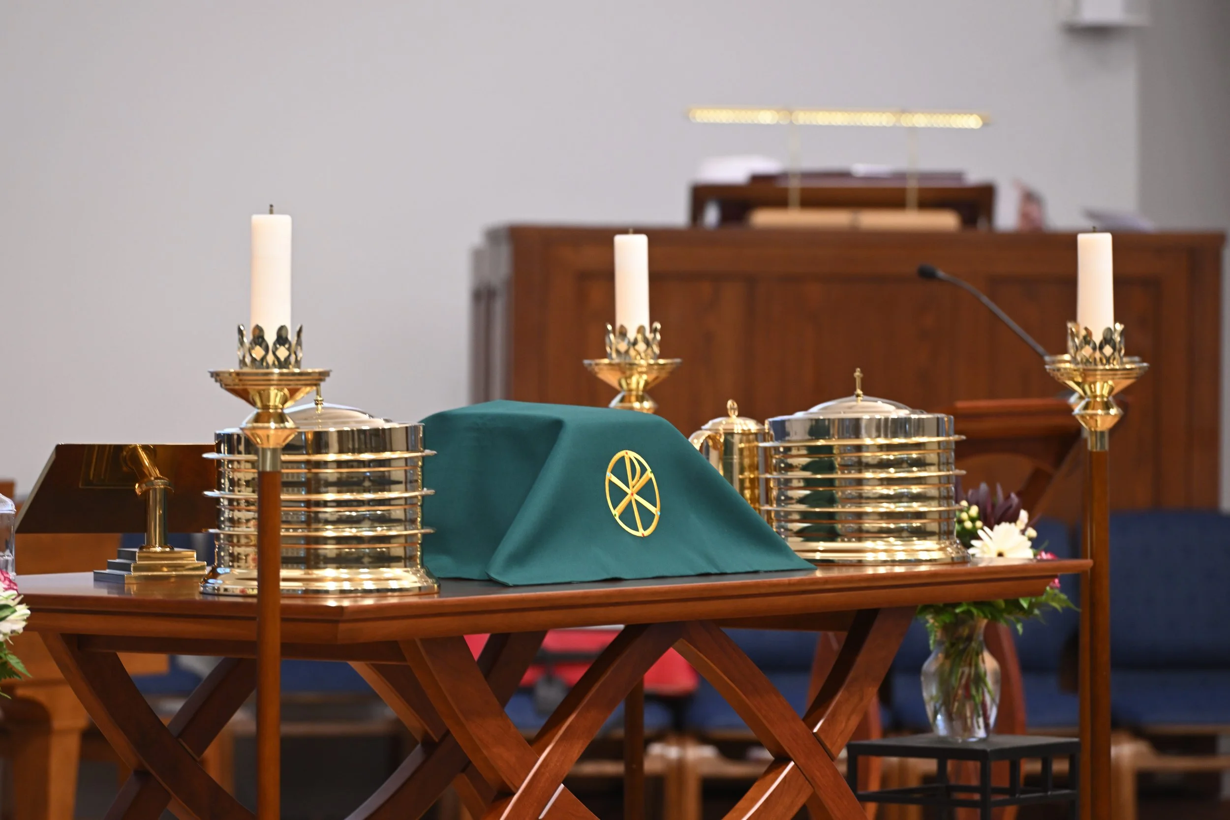 A church altar with three lit candles in gold holders, a green cloth with a gold emblem, and a cross symbol, surrounded by flowers and greenery in a vase, with a wooden pulpit in the background.
