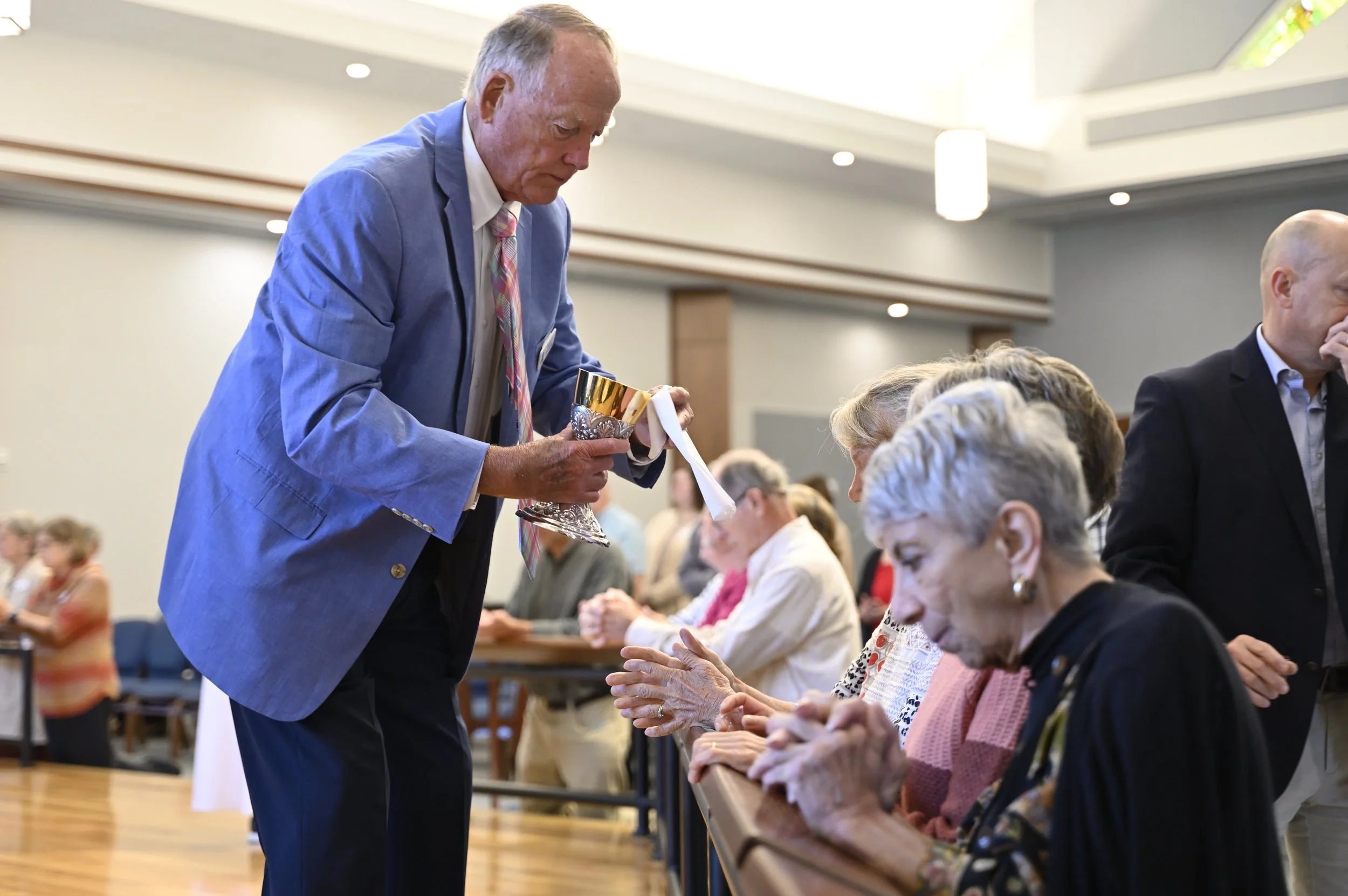 An elderly man in a blue suit and colorful tie is holding a chalice and a towel while offering communion to seated elderly women during a religious service. The women are receiving communion with their hands extended, seated at a long table. The setting appears to be a church or community center.
