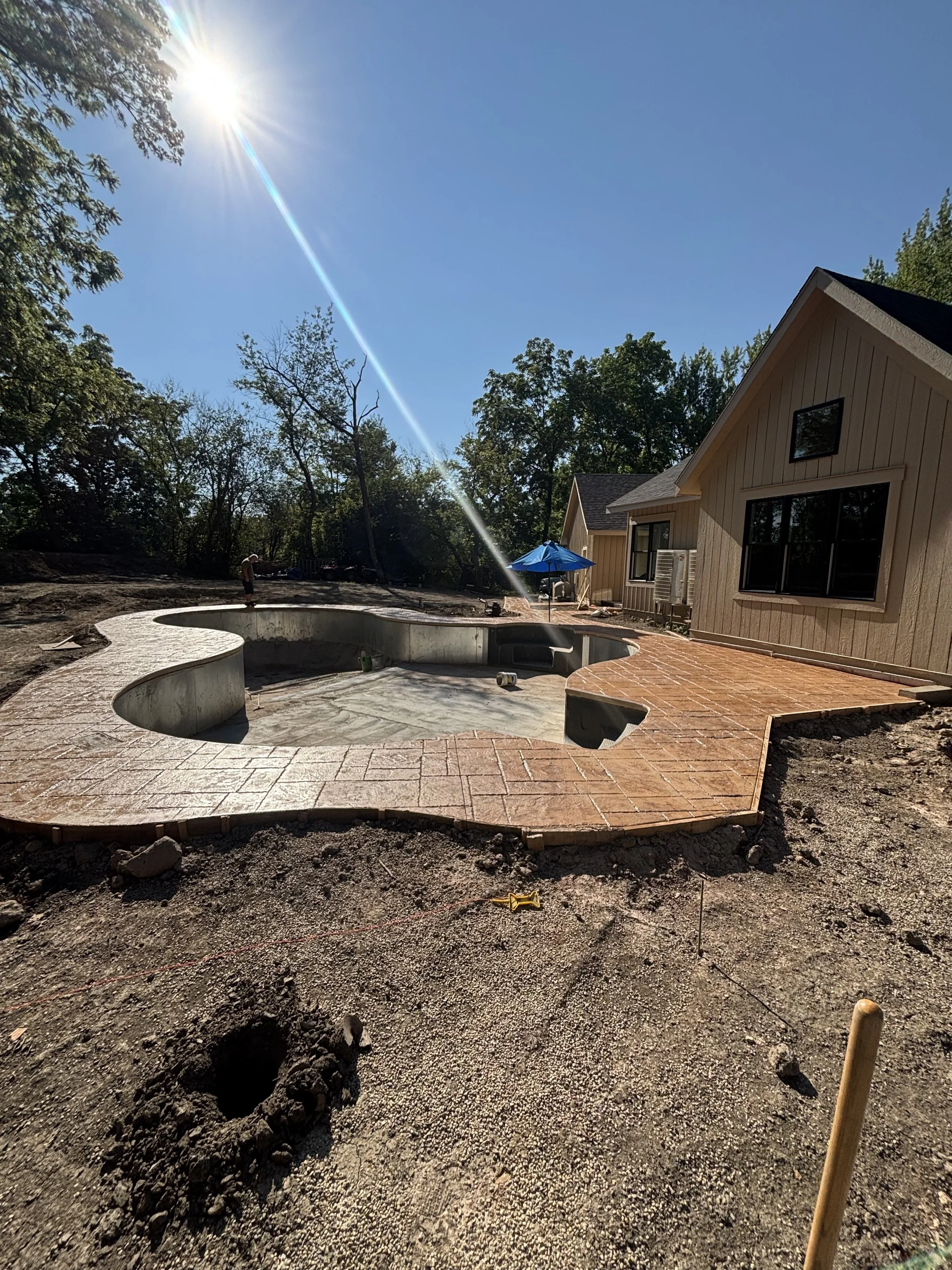 Construction site with a circular concrete structure, possibly a pool, surrounded by brick paving, next to a house with beige siding and black windows, trees in the background, sunny sky, and an umbrella.
