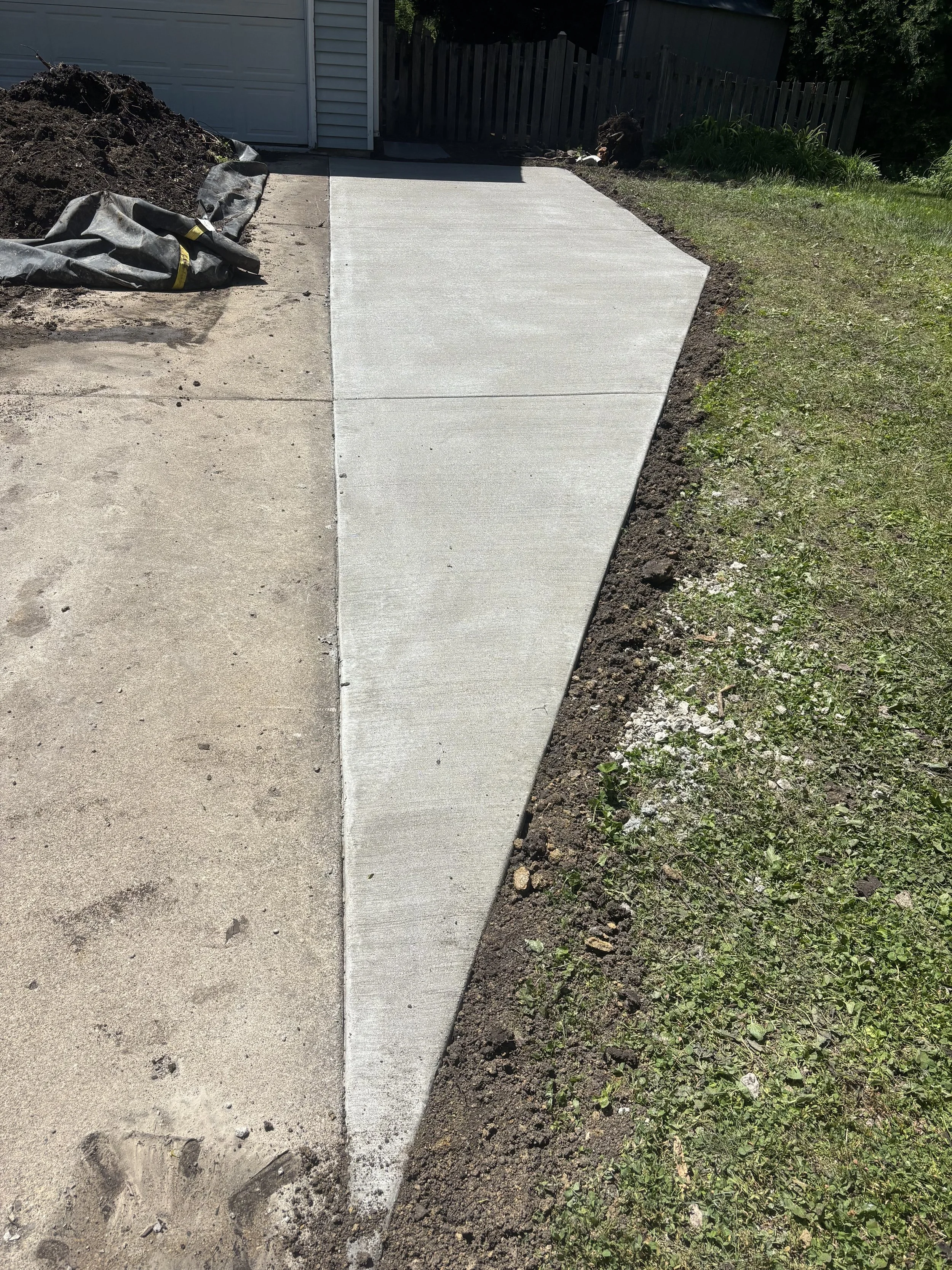 Newly poured concrete sidewalk next to grass, with a wooden fence and house garage in the background.