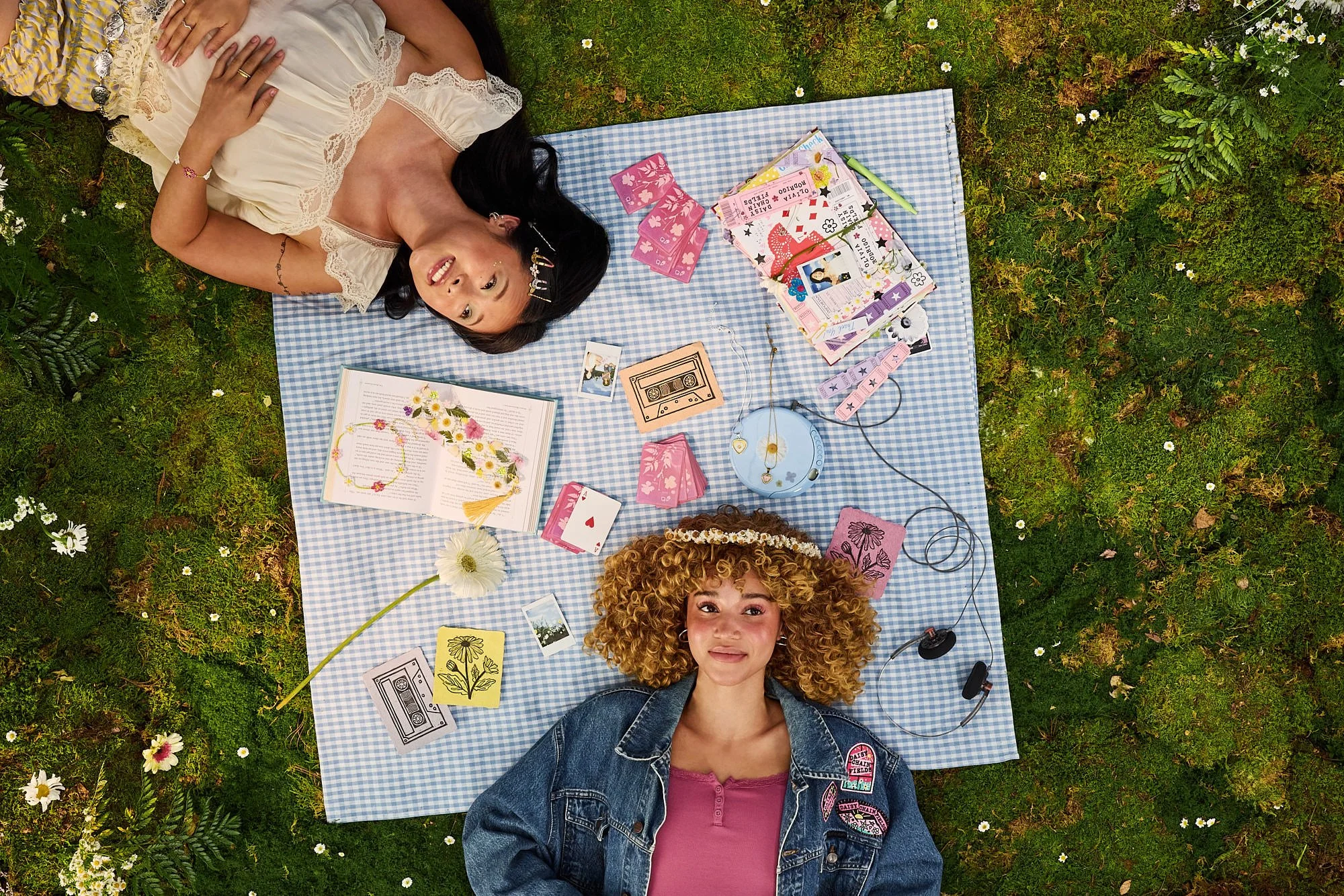 Three women lying on a blue checkered picnic blanket in a grassy area with wildflowers. They have various items around them, including books, photographs, cards, jewelry, and small decorative objects. They are smiling and appear to be enjoying a rela