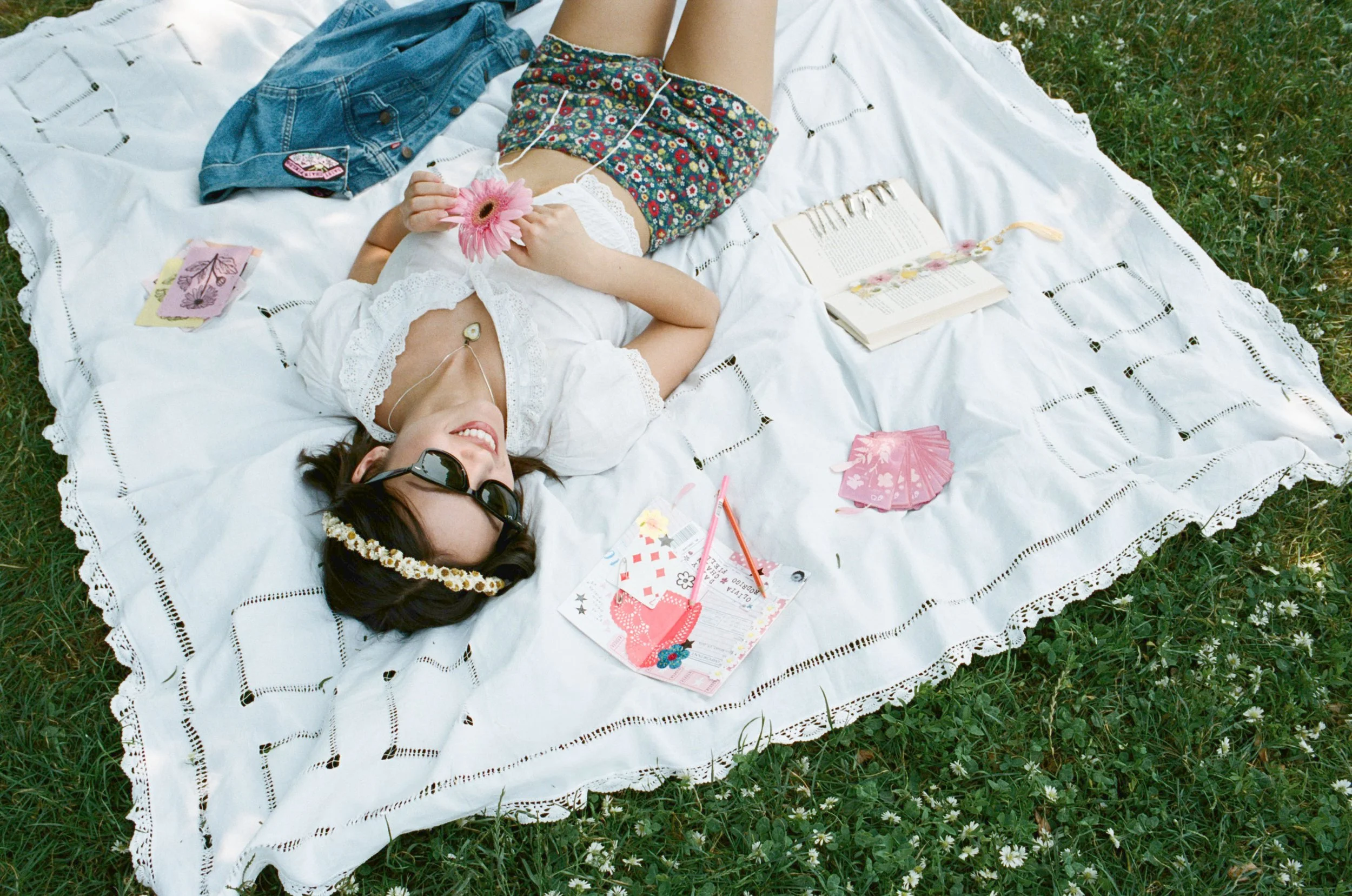 A woman laying on a white blanket with black embroidered designs on the grass, holding a pink flower. She is smiling, wearing sunglasses, a white lace blouse, and floral shorts. There are books, colorful cards, a pink parasol, and a pink and white pa