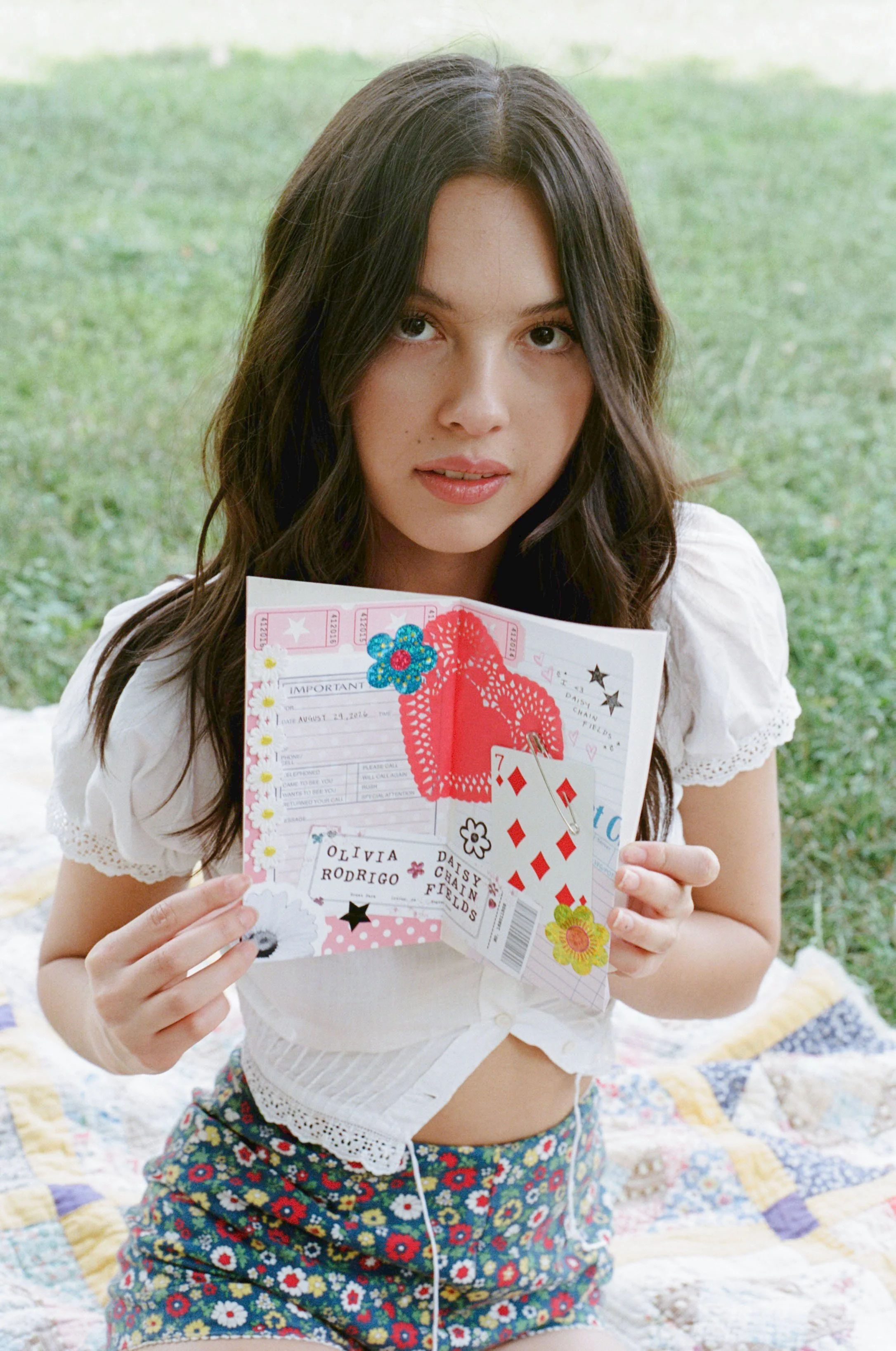 A young woman with long, dark hair holding a colorful invitation or card with floral and heart designs. She is sitting on a quilt outside on grass, wearing a white top with lace details and floral shorts.