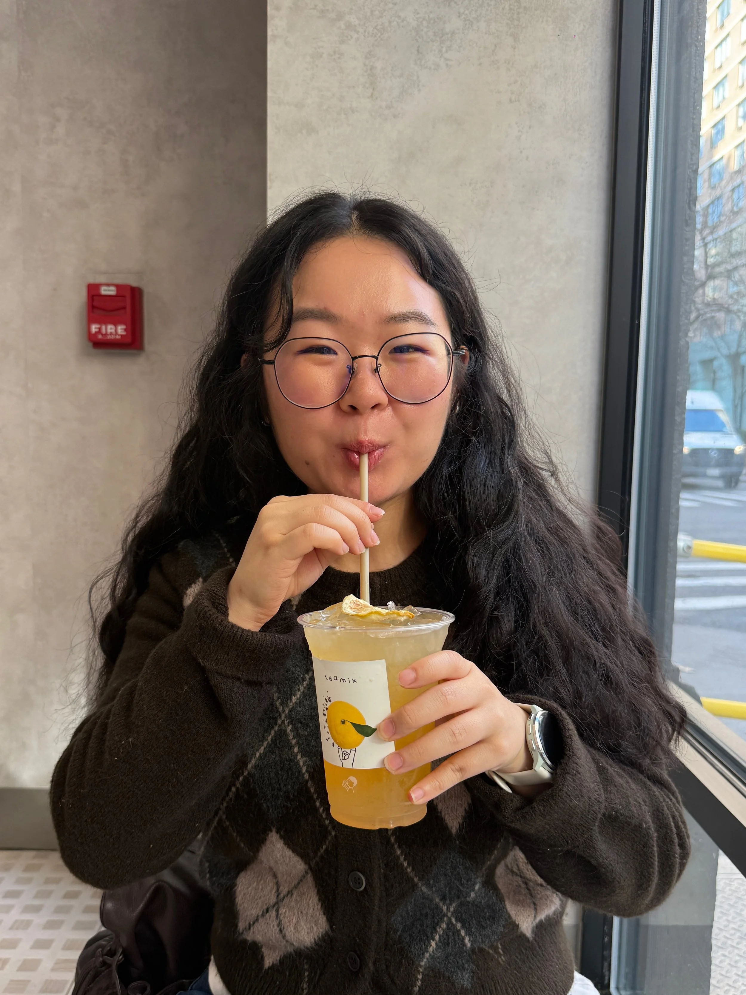 A woman with glasses and long dark hair, smiling slightly, is sitting by a window and drinking a yellow-colored beverage from a clear plastic cup with a paper sleeve, using a straw.