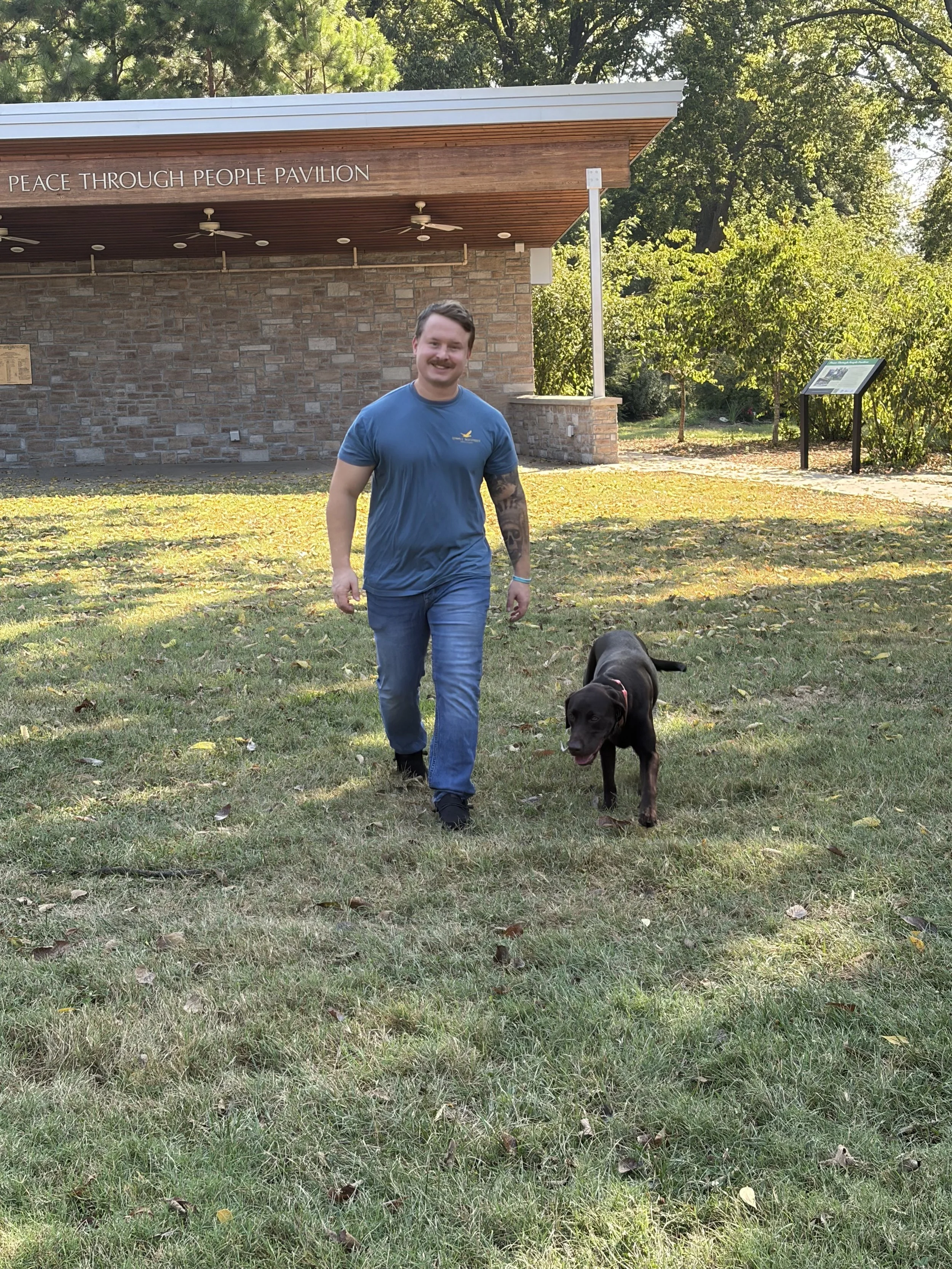 A smiling man wearing a blue T-shirt and jeans walking a black dog on a grassy area near a pavilion with a sign that reads 'Peace Through People Pavilion' and surrounded by trees.