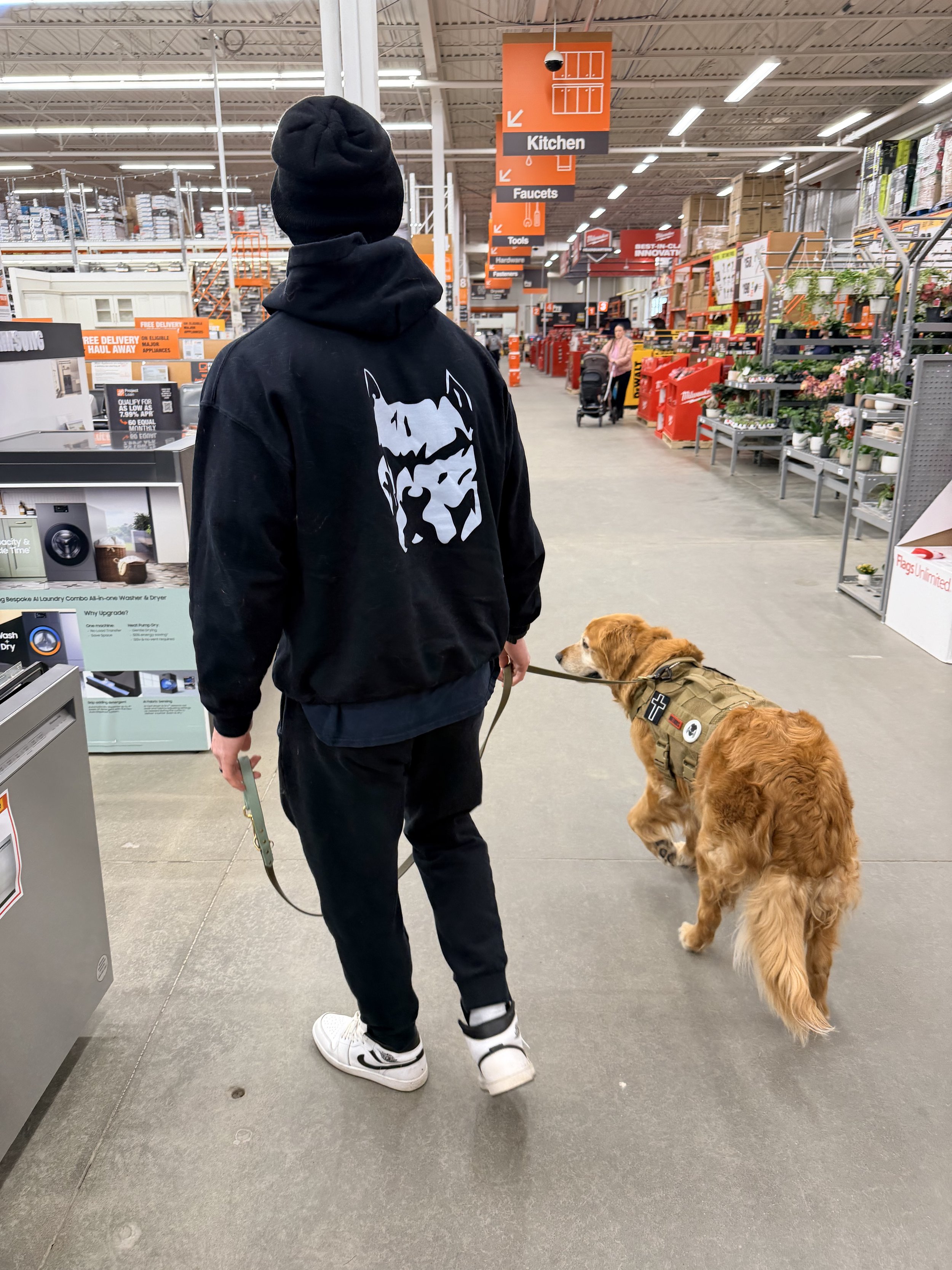 A person with black clothing and a black beanie walking a golden retriever dog inside a home improvement store. The store has orange signage, aisles with various tools and plants, and a woman pushing a shopping cart in the background.