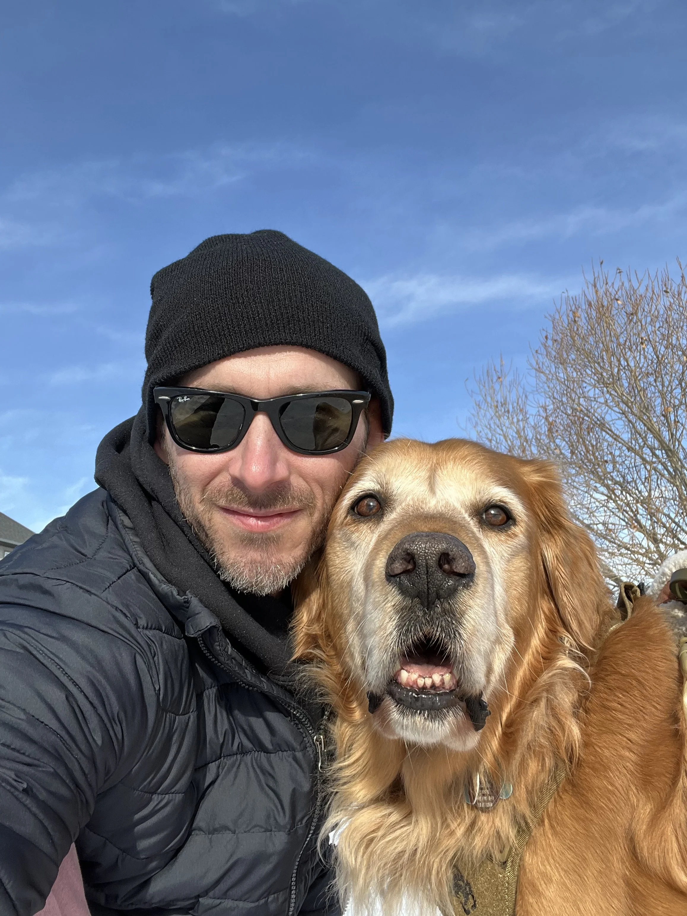 A dog trainer wearing sunglasses, a black beanie, and a black jacket taking a selfie with a golden retriever outdoors against a bright blue sky and leafless tree.