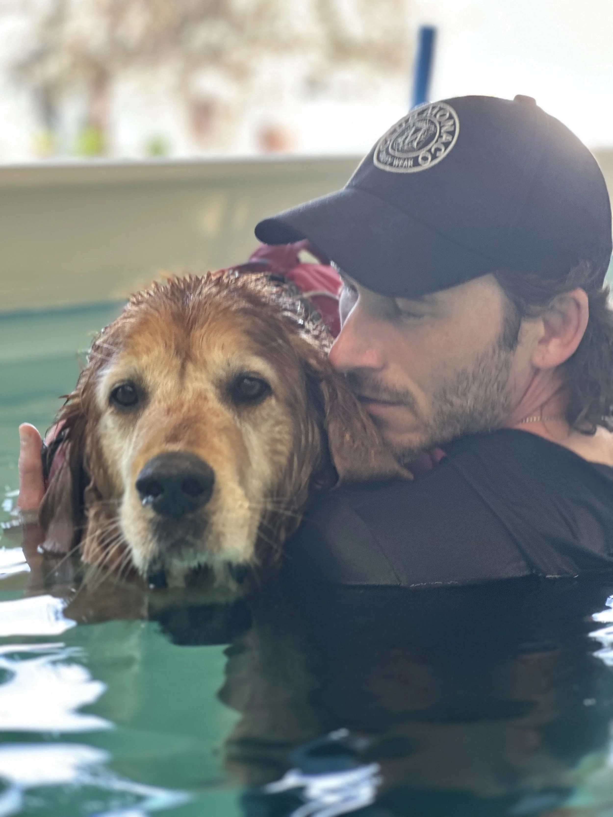 A man wearing a dark cap with a circular logo embraces a wet golden retriever dog in a pool or body of water.