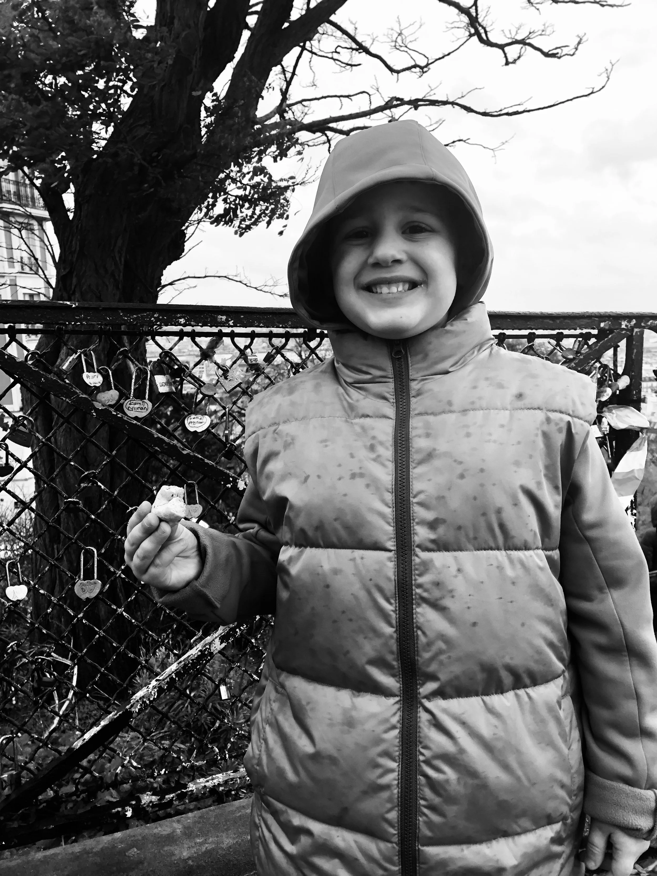 A smiling young girl in a hooded jacket holding a small object in her hand, standing in front of a chain-link fence with many padlocks attached, a large leafless tree, and a cloudy sky in the background.
