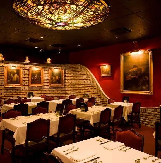 Elegant restaurant dining room with brick wall, framed paintings, and a decorative ceiling light fixture.