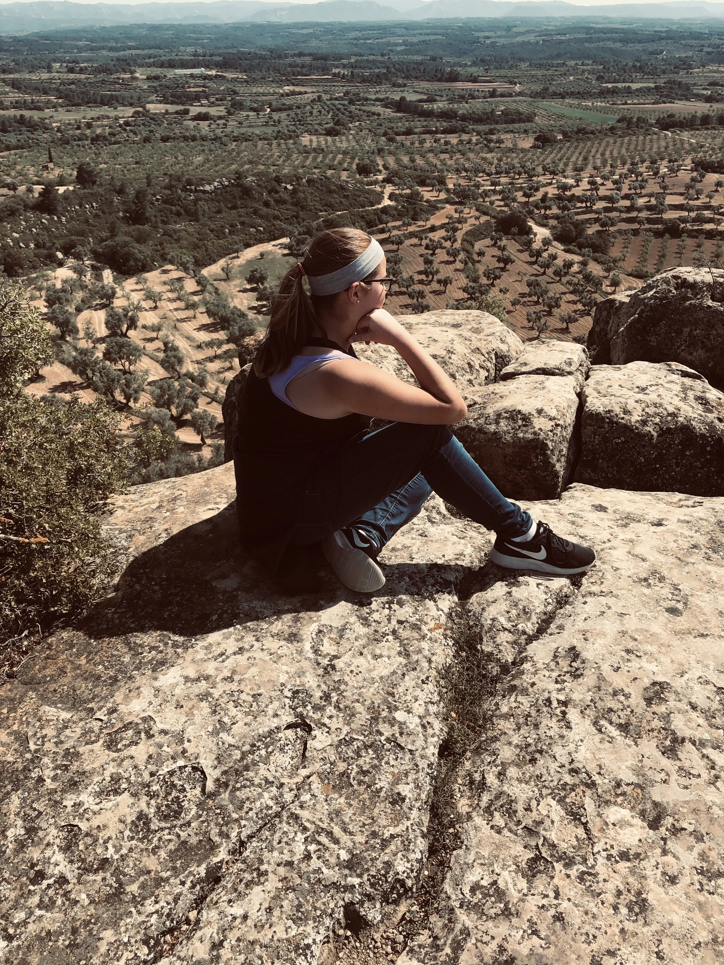 A young woman wearing glasses, a gray headband, a black top, and athletic shoes is sitting on large rocks, looking out over a vast landscape of farmland with rows of trees and fields, under a hazy sky.