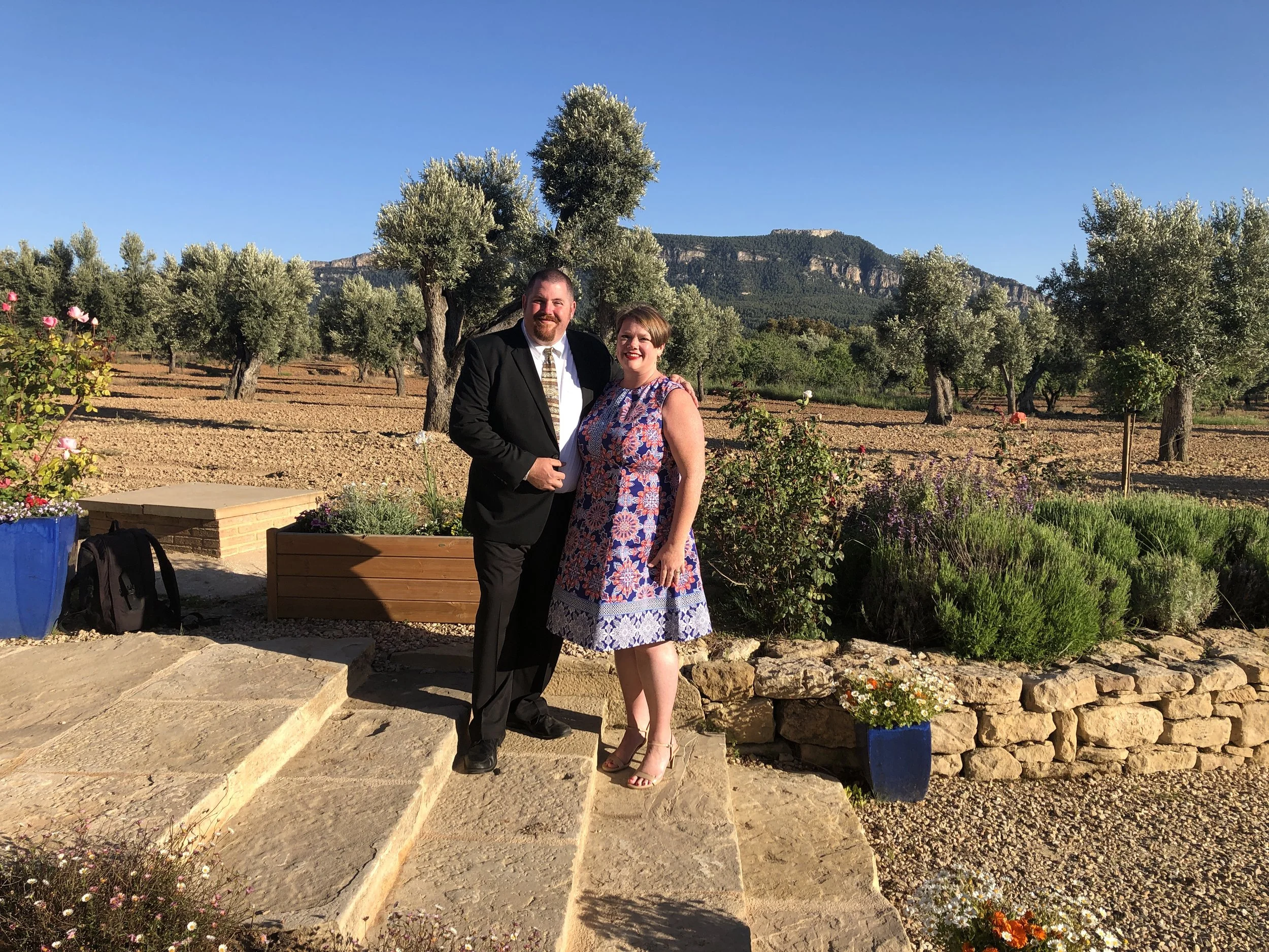 A man in a black suit and a woman in a floral dress smiling and standing outside in a garden with trees, flowers, and mountains in the background.