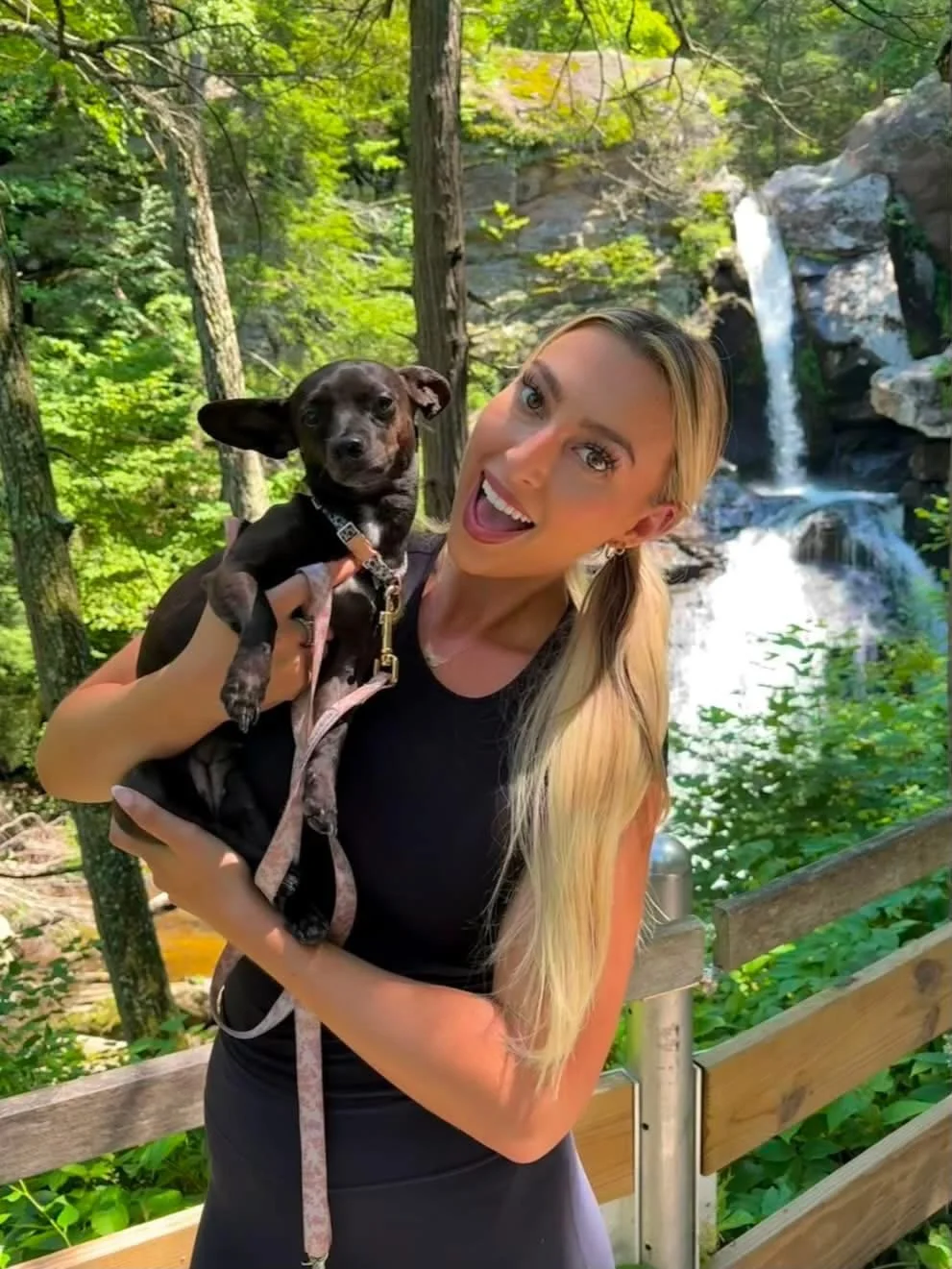 A woman holds a small black dog with one ear standing up, in front of a waterfall in a lush green forest.