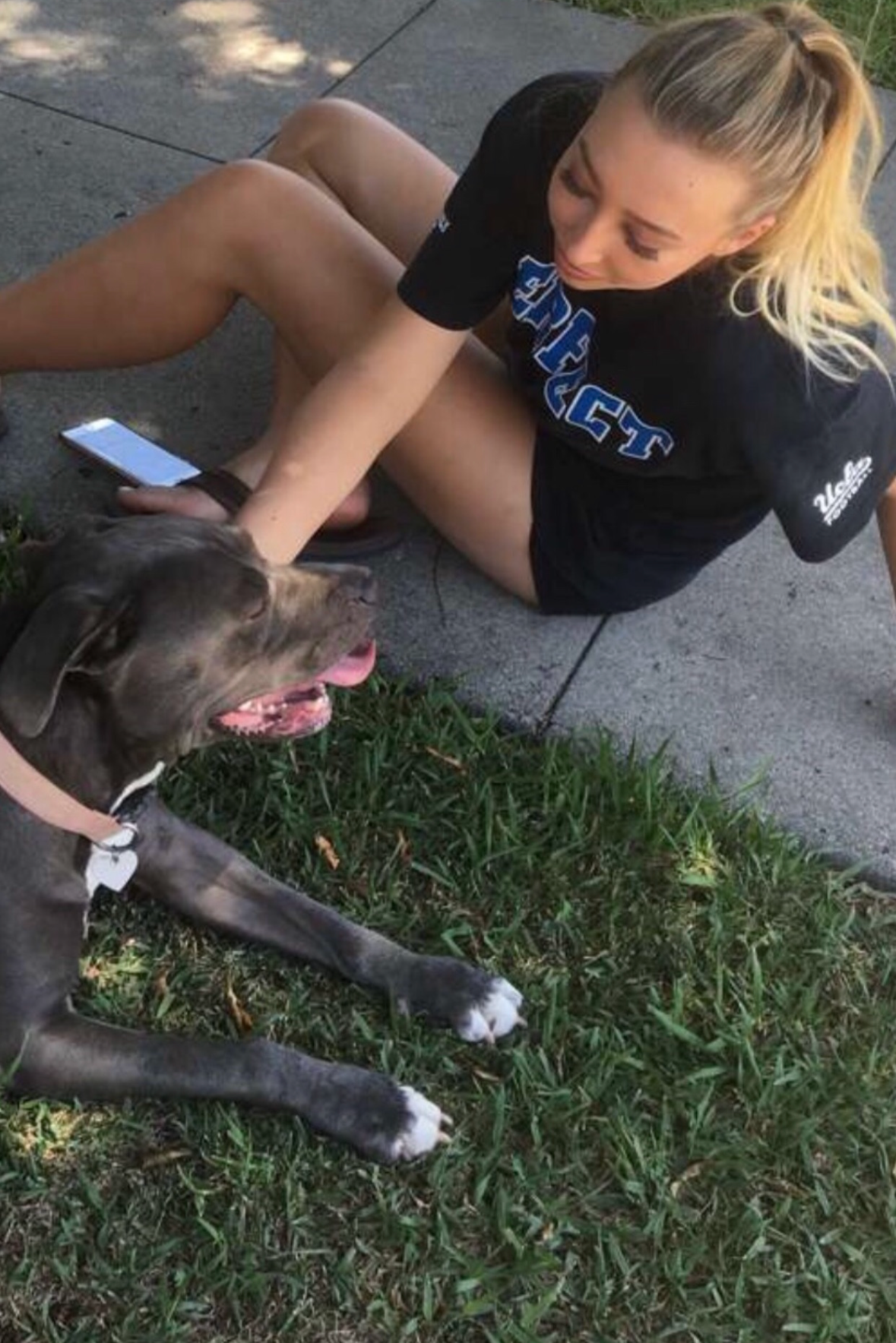 A woman sitting on a concrete sidewalk outdoors with a dog lying beside her on the grass. The woman has blonde hair tied back and is wearing a black t-shirt and shorts. The dog is a large breed with gray and white fur, lying with its tongue out.
