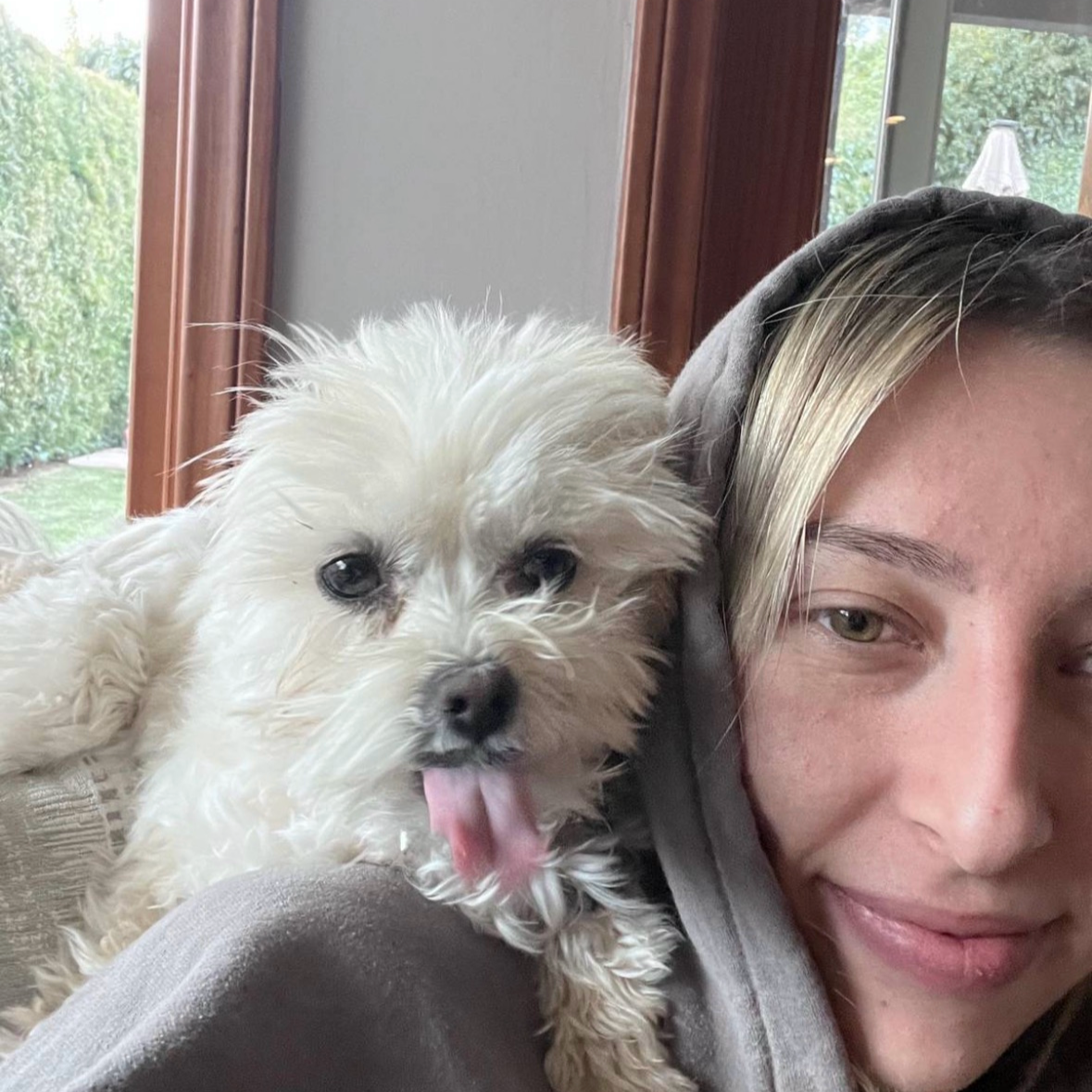 A person with blonde hair and a gray hoodie close to a small white dog with curly fur, both lying indoors near a window with trees outside.