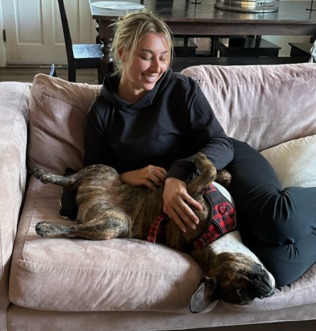 A woman with blonde hair in a black hoodie smiling while lying on a pink couch with a brindle dog wearing a red harness, who is lying on its back with paws in the air.