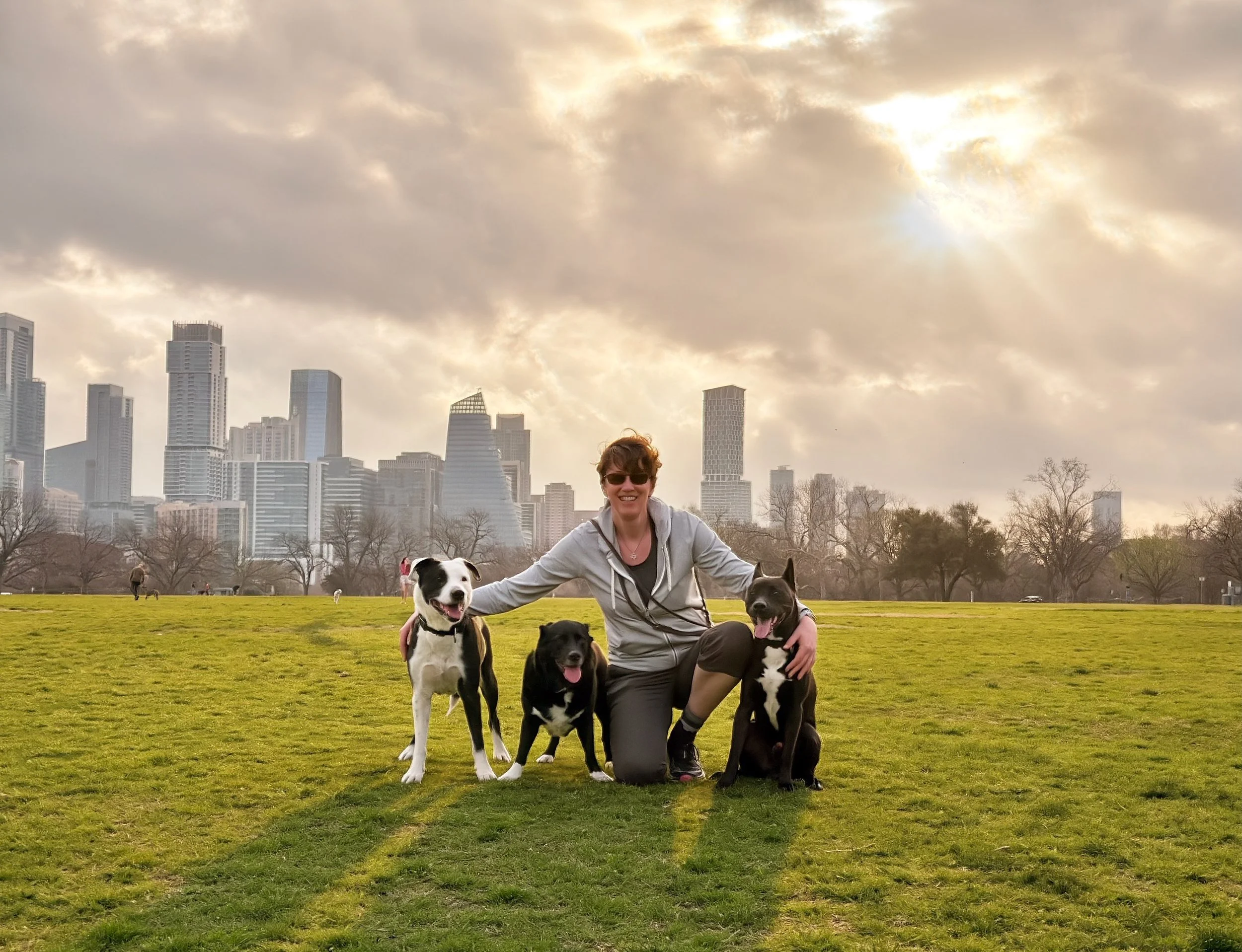 A woman smiling and wearing sunglasses, crouching on a grassy field surrounded by four black and white dogs, with a city skyline and cloudy sky in the background.
