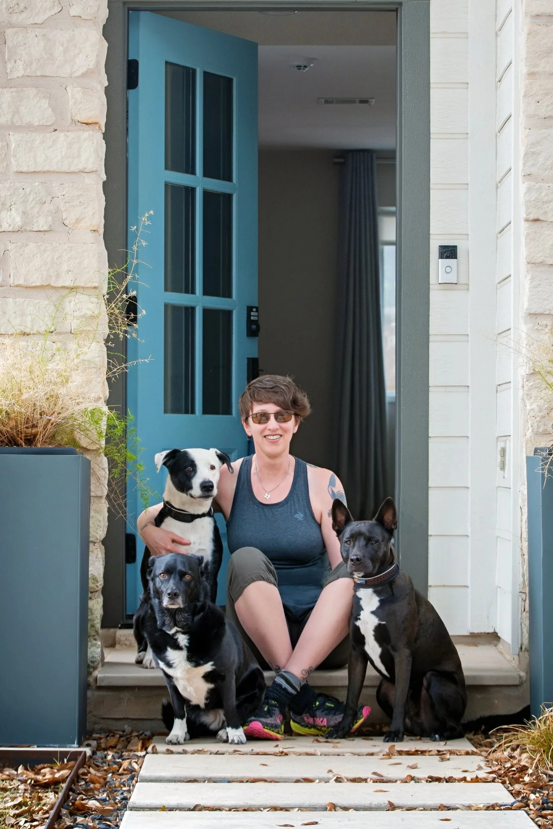 A person sitting on the front porch step with four dogs, smiling, in front of a house with a blue door and stone exterior wall.