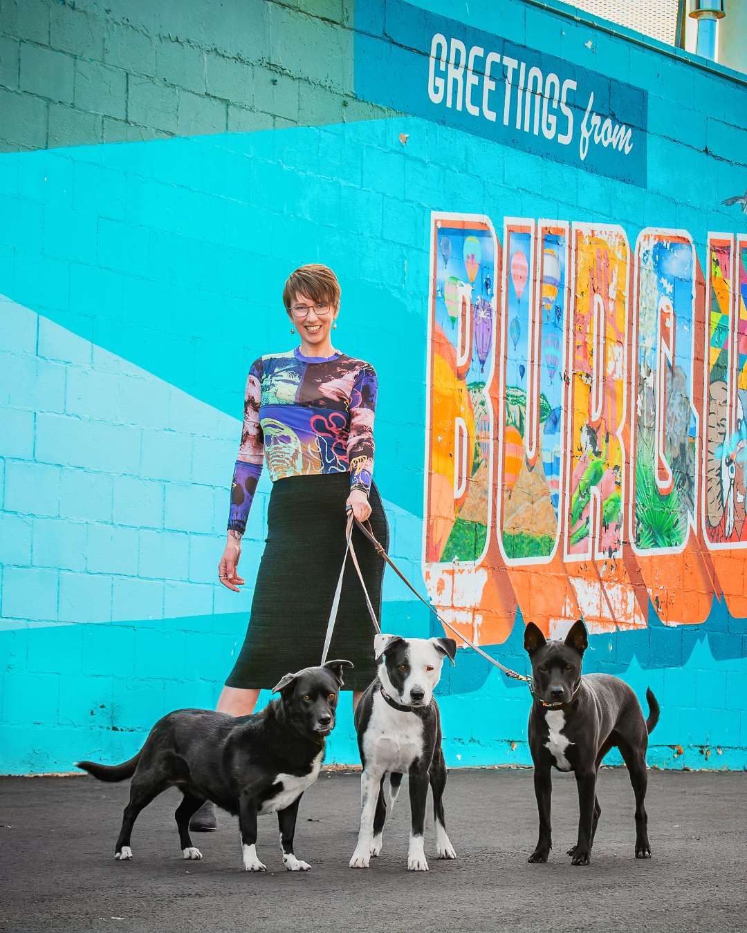 A woman standing on a street holding three dogs on leashes in front of a colorful mural with the words "Greetings from San Jose" painted on a brick wall.