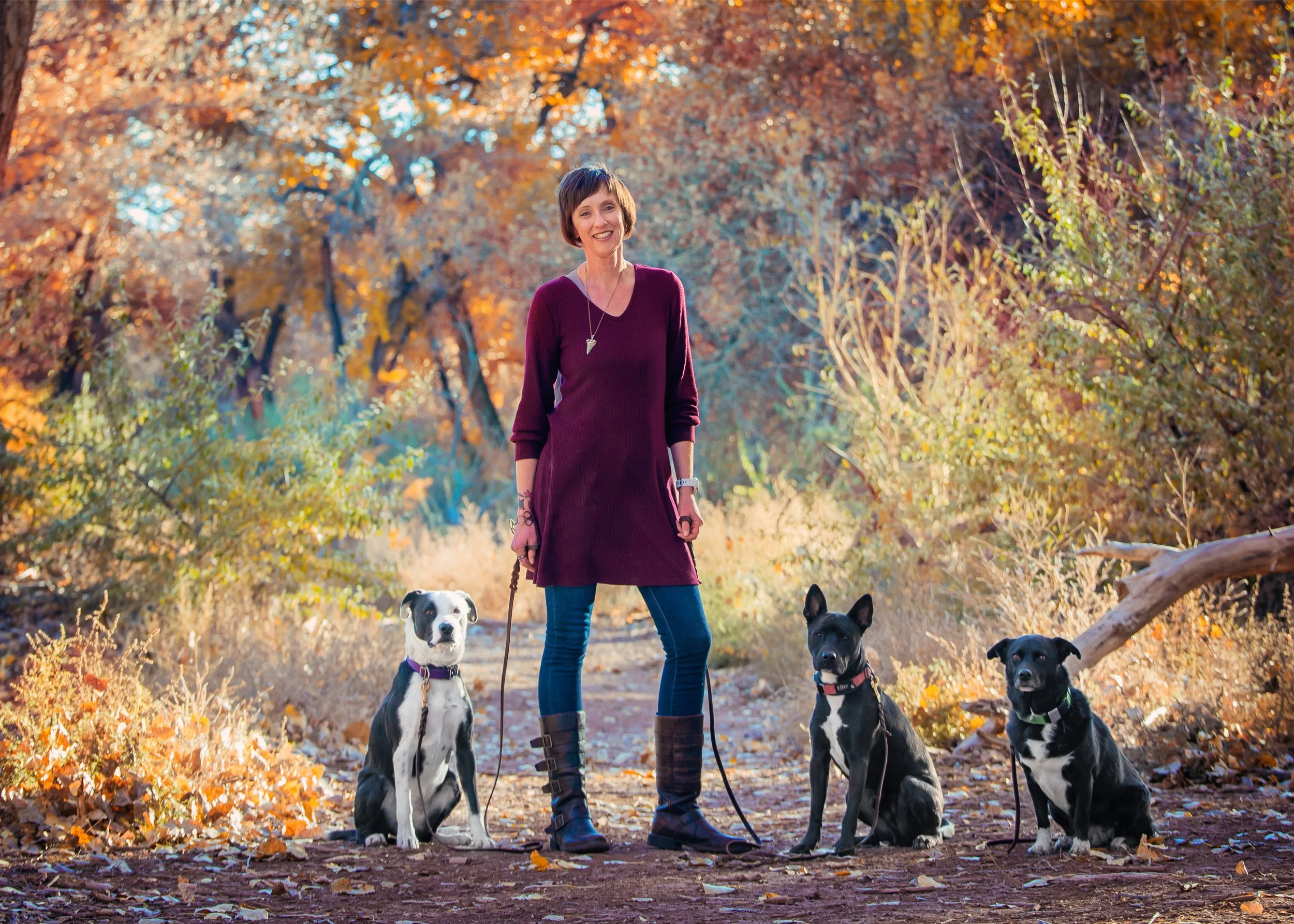 A woman with short brown hair wearing a maroon dress standing outdoors in an autumn forest with four dogs on leashes, surrounded by fall foliage with orange, yellow, and brown leaves.