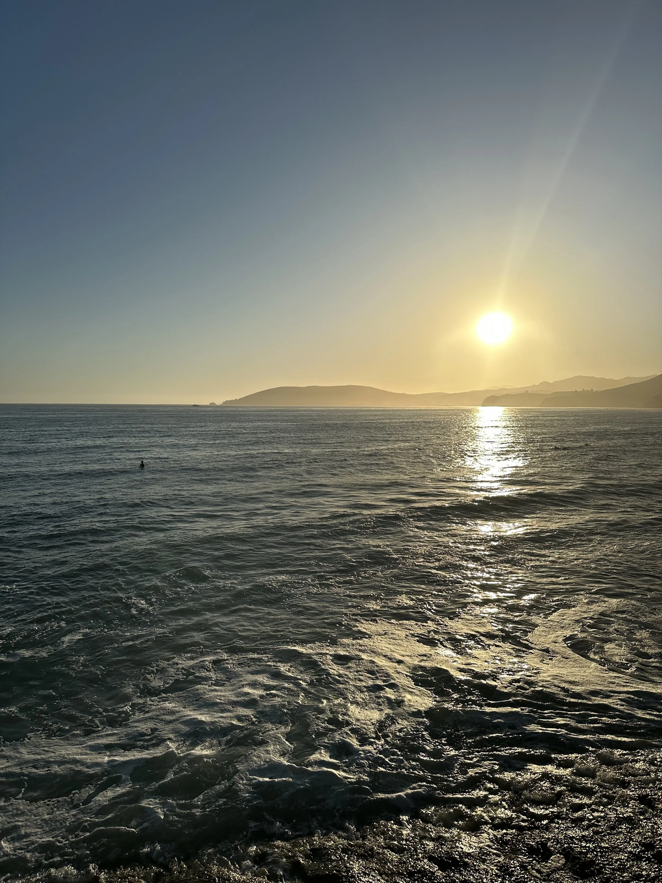 Sunset over the ocean with distant land and mountains, gentle waves on the water, and a clear sky.