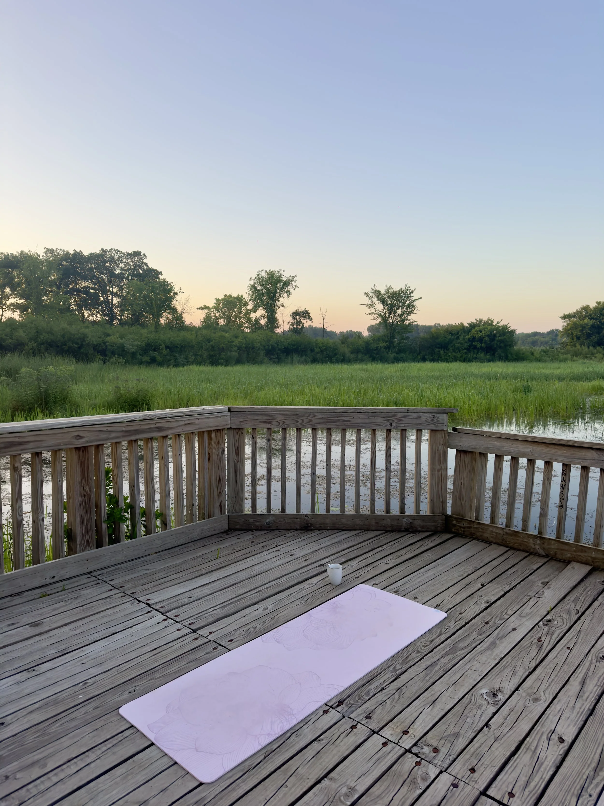 A wooden deck overlooking a green field with trees in the background and a clear sky during sunset. There is a white cup on the deck and a sheet of paper with a drawing on it.