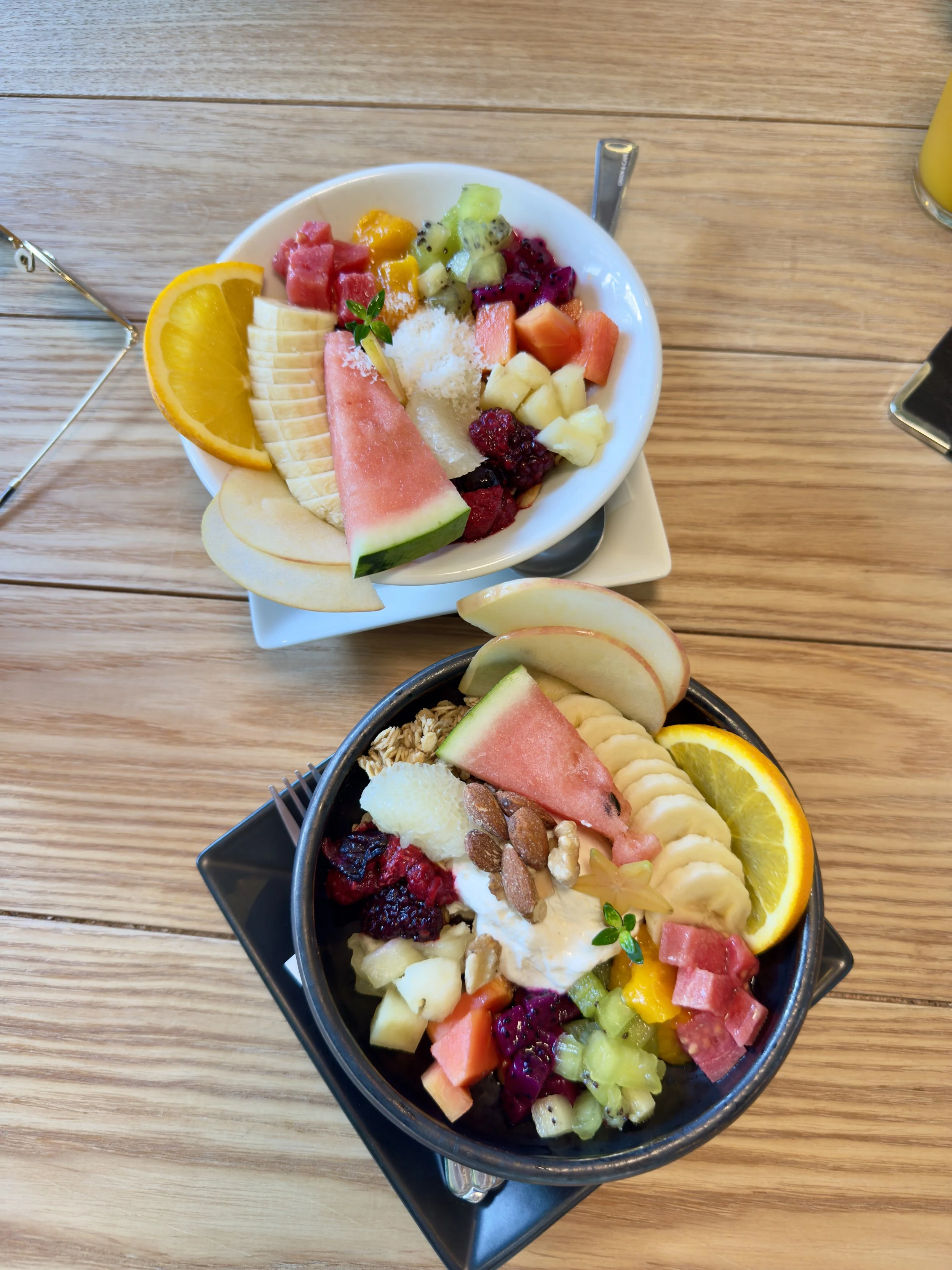 Two bowls of colorful fruit salad on a wooden table, garnished with orange slices, melon, pineapple, watermelon, berries, and topped with nuts and herbs.