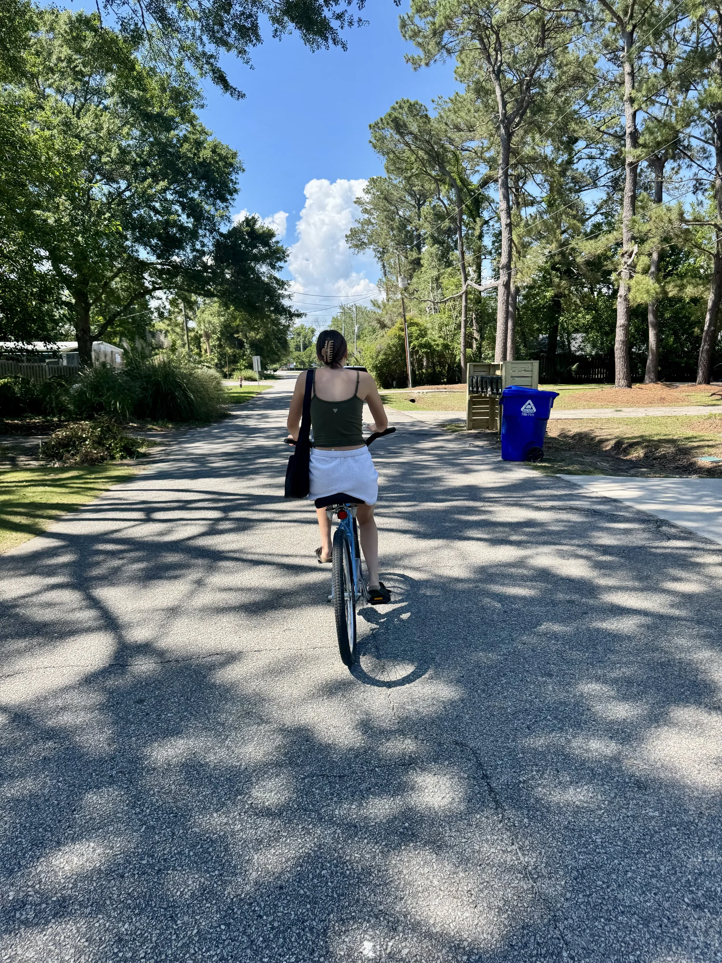 A person riding a bicycle on a shaded residential street under a clear blue sky, with trees, a mailbox, and a blue recycling bin visible.