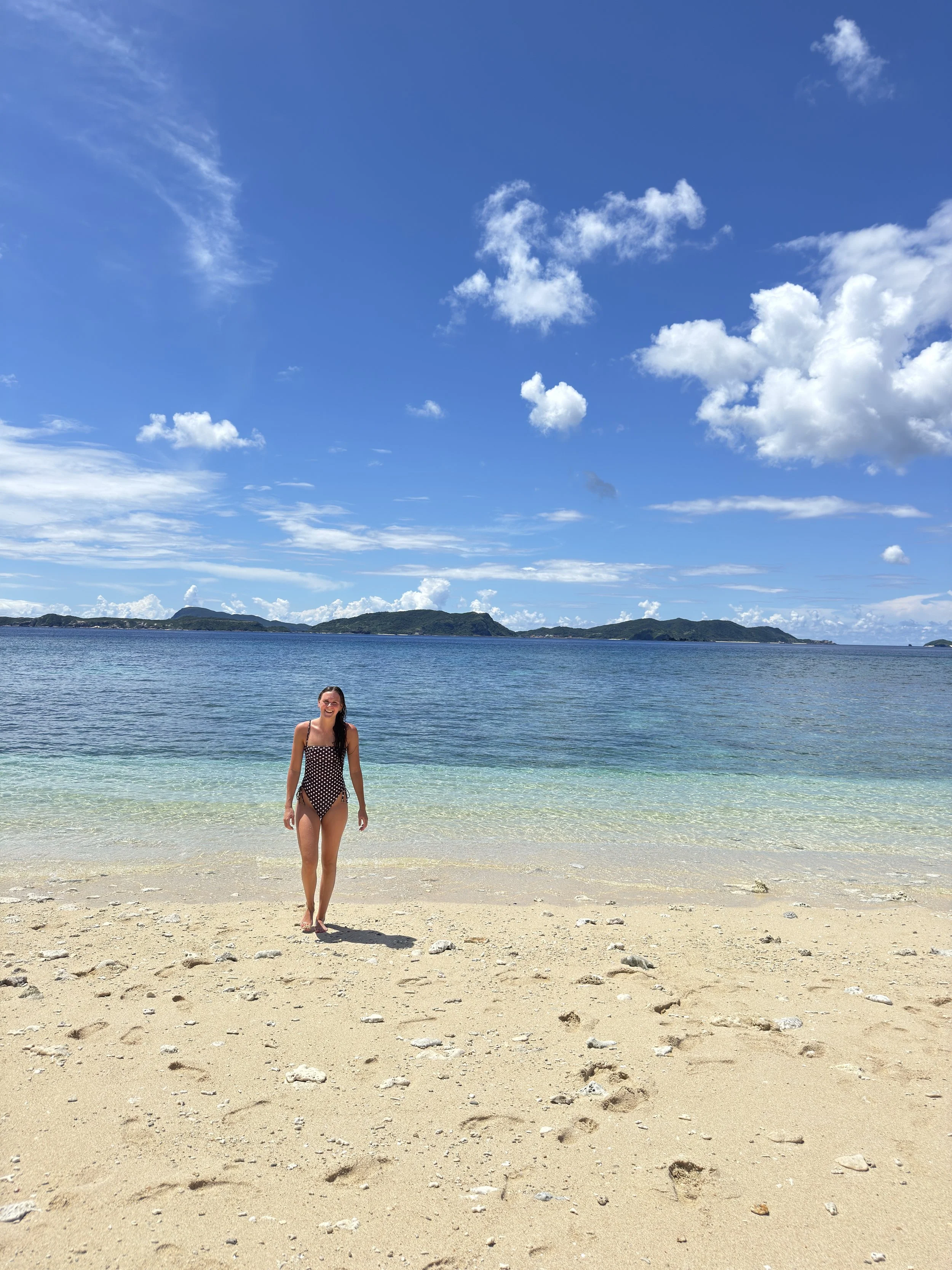 A woman in a polka-dot swimsuit standing on a sandy beach with clear water, a distant green island, and a bright blue sky with white clouds.