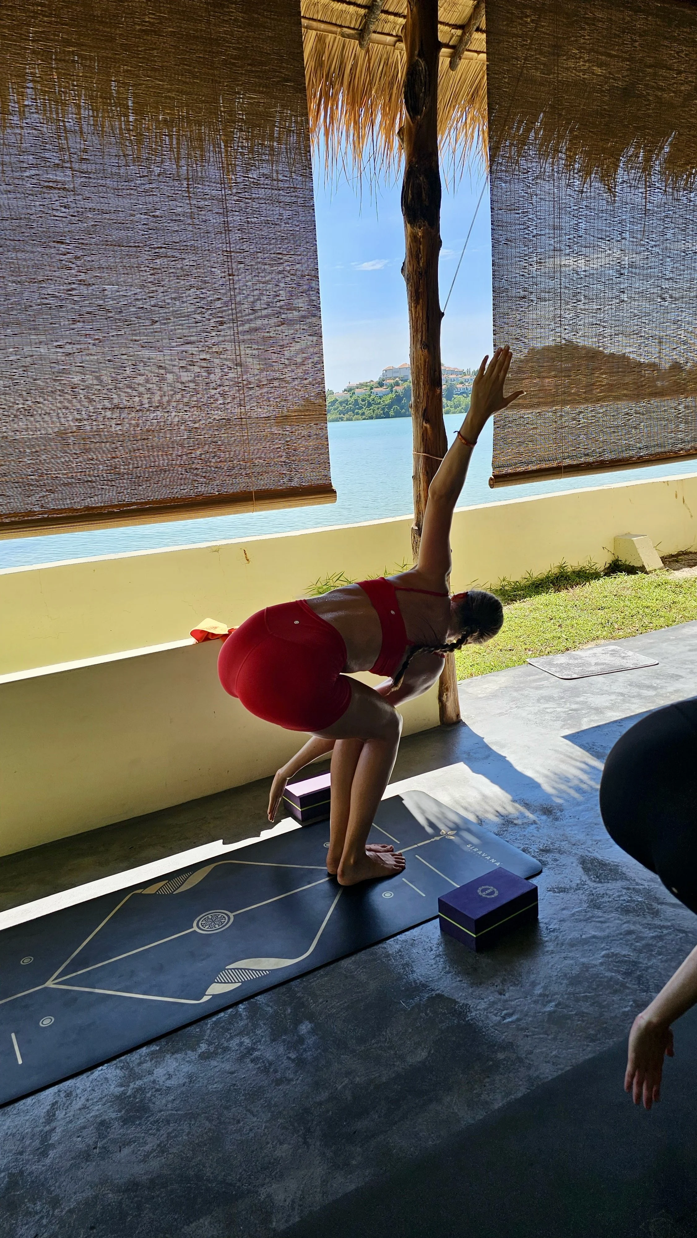 A woman wearing red athletic clothing is practicing yoga outdoors beneath a thatched roof with bamboo blinds, near water. She is in a forward bend pose, with one hand reaching toward the ceiling and the other on the ground, on a yoga mat with a geometric design. There are yoga blocks on the ground nearby.