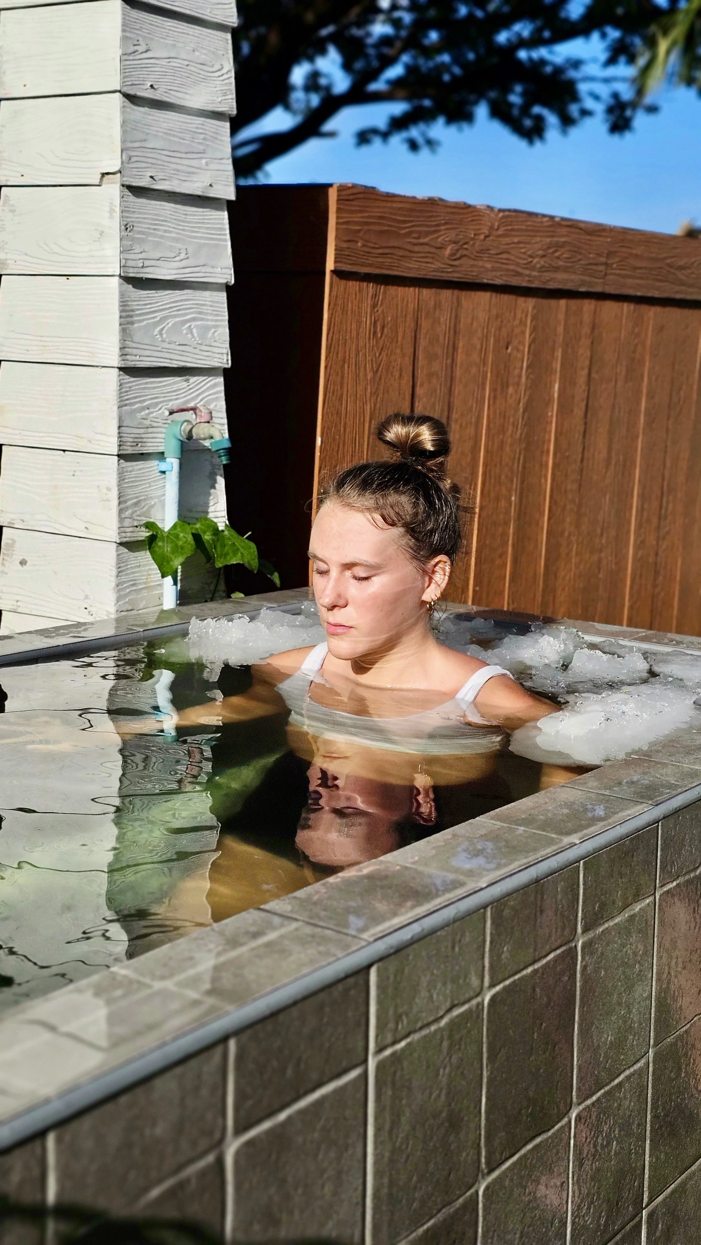 Woman relaxing in an outdoor bathtub surrounded by a wooden fence with a blue sky and tree in the background.