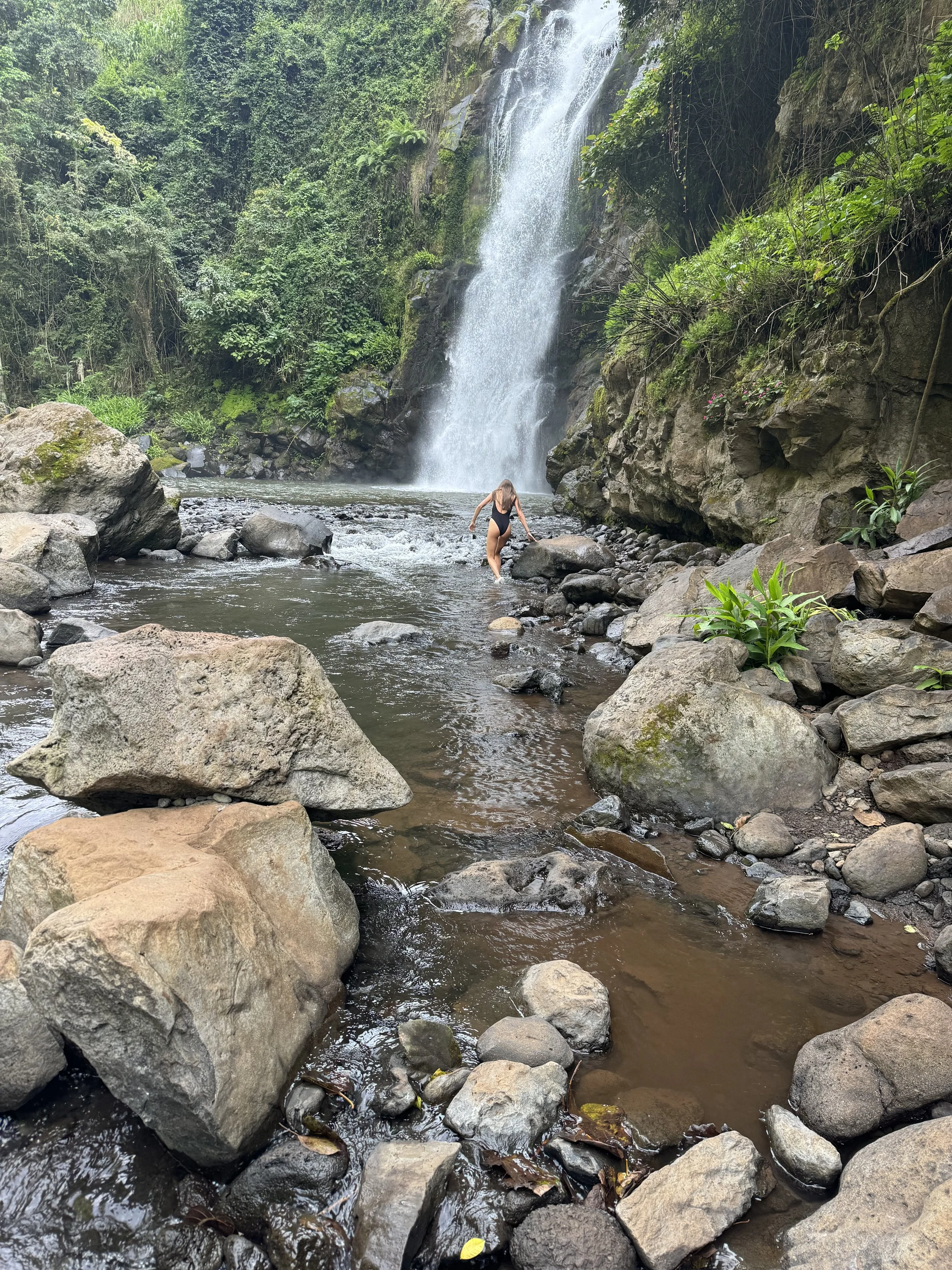 A woman in a black swimsuit walking on rocks in a river beneath a waterfall in a lush green forest.