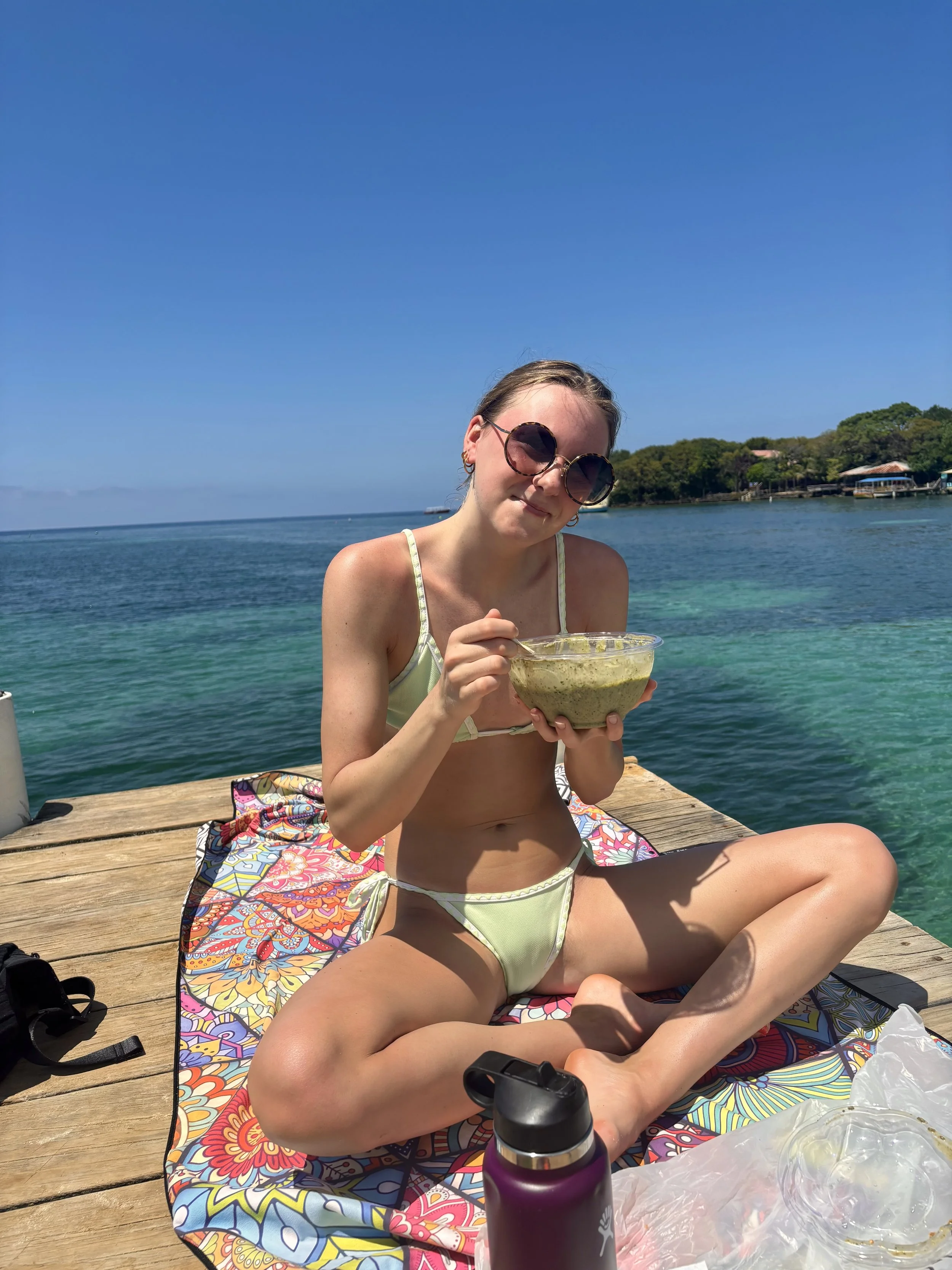 Young woman sitting on a wooden dock by the water, holding a bowl of guacamole, wearing sunglasses and a bikini, with the ocean and trees in the background.