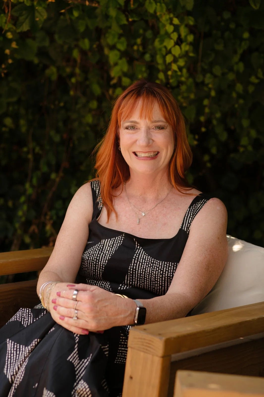 A woman with red hair wearing a black and white patterned sleeveless dress, sitting on a wooden bench outdoors, smiling with her hands clasped, against a background of green foliage.