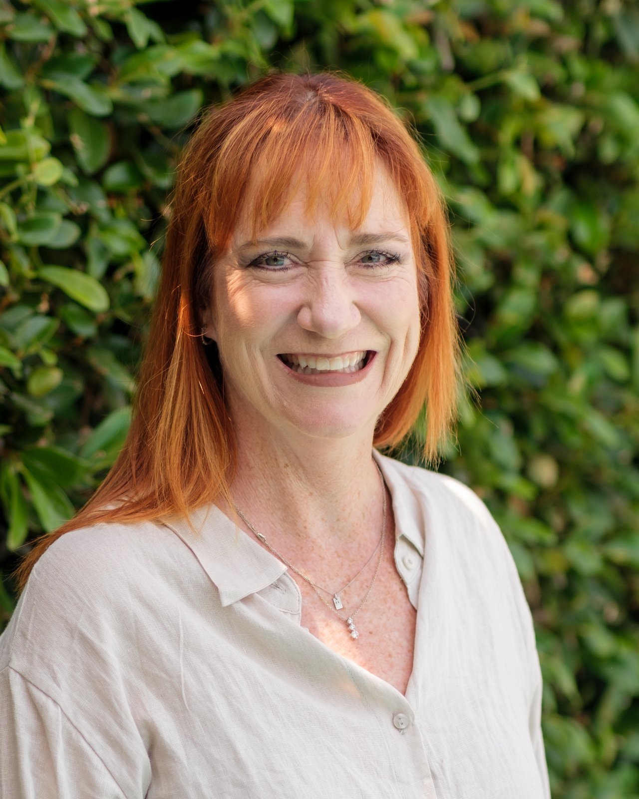 A woman with red hair smiling outdoors in front of green foliage.