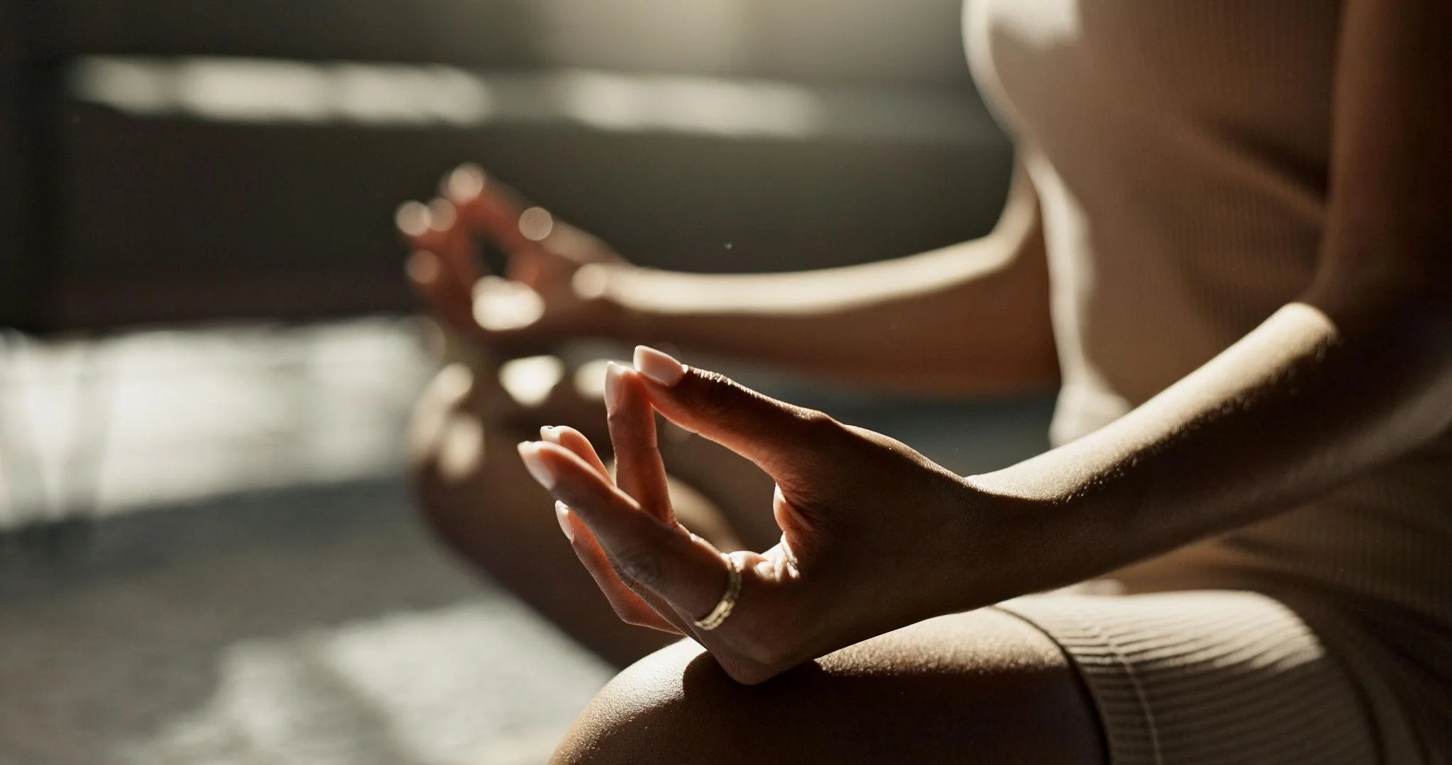 Close-up of a person's hand with fingers in a mudra gesture, sitting in a meditative pose, with natural sunlight streaming in.