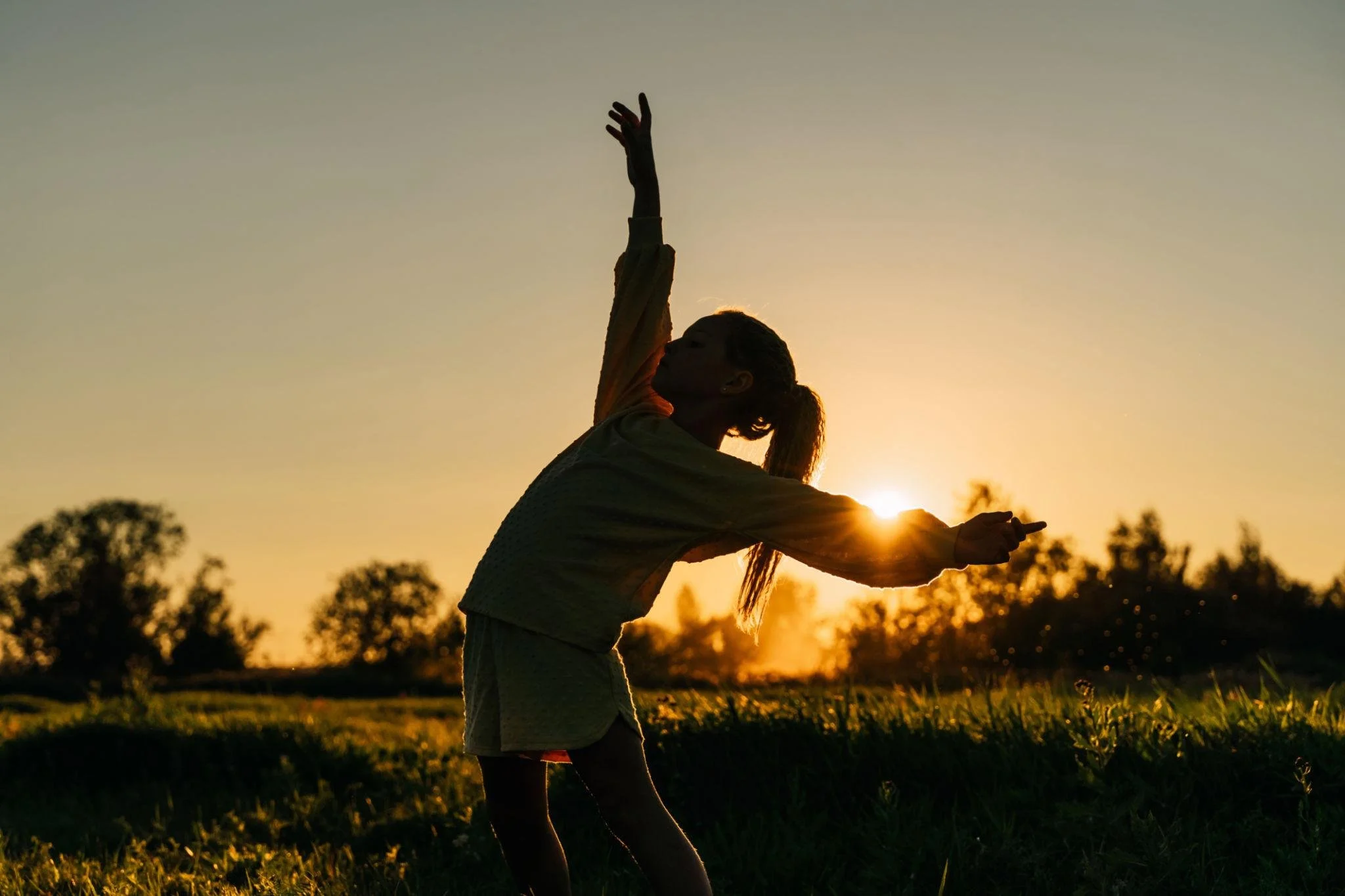 A woman dancing or exercising outdoors at sunset, raising one arm upward, with trees and a field in the background.