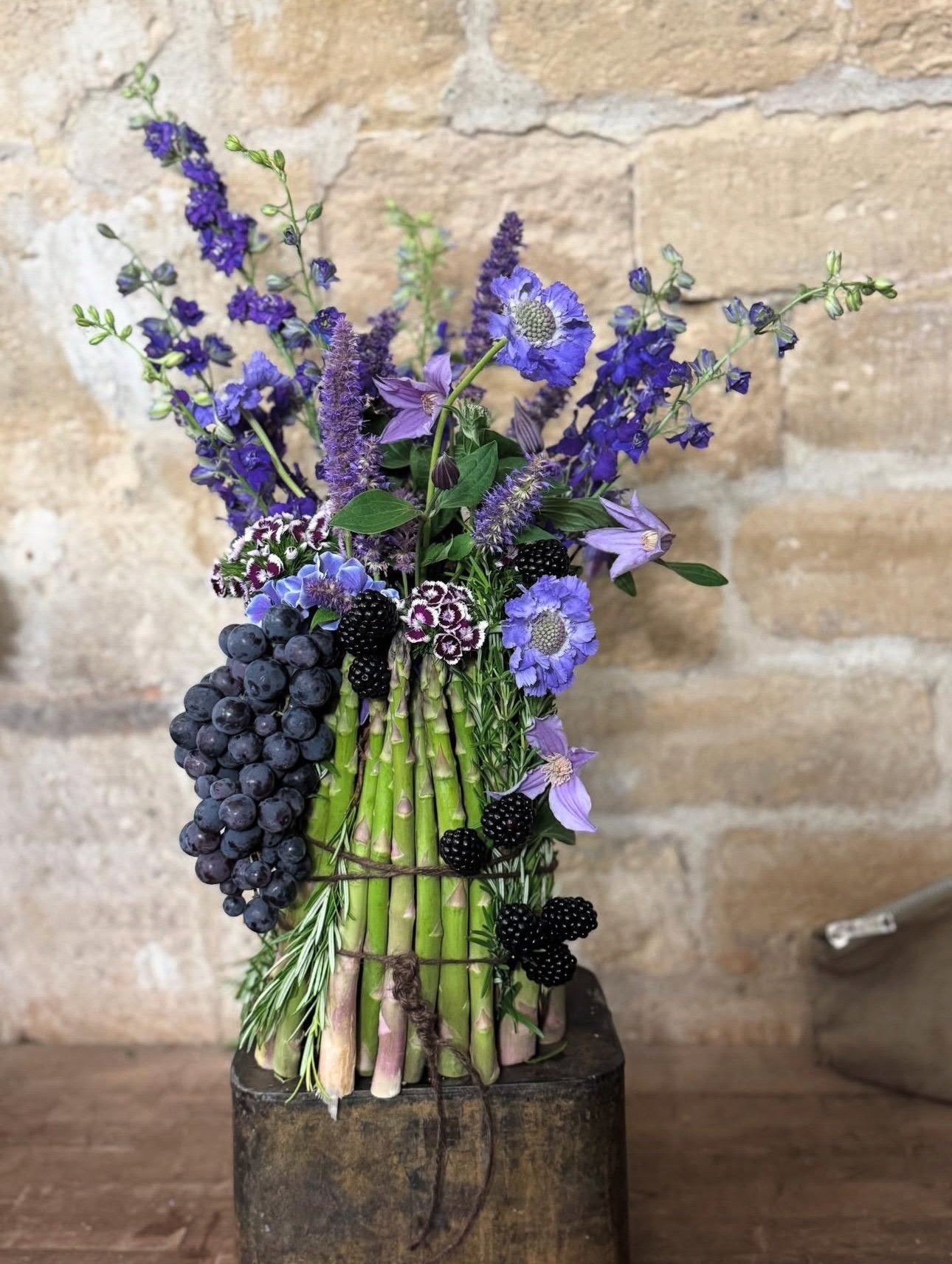 A bouquet of purple flowers, blackberries, and grapes arranged in a vase made of cut green stems, set against a brick wall background.
