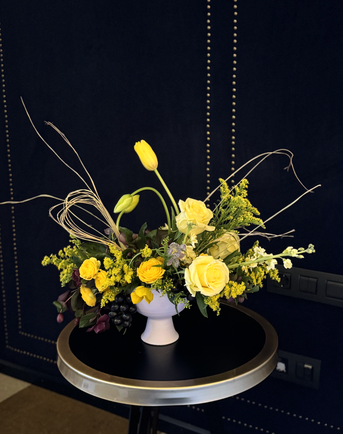 A floral arrangement with yellow roses, tulips, and other yellow and purple flowers in a white vase, placed on a black round table against a dark blue wall.