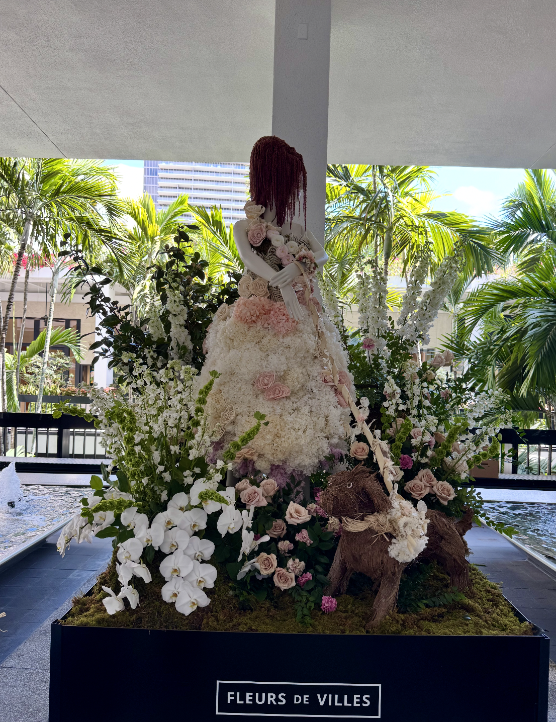 A decorative floral sculpture of a woman in a floral dress surrounded by white, pink, and purple flowers with green foliage, set outdoors with palm trees and city buildings in the background.