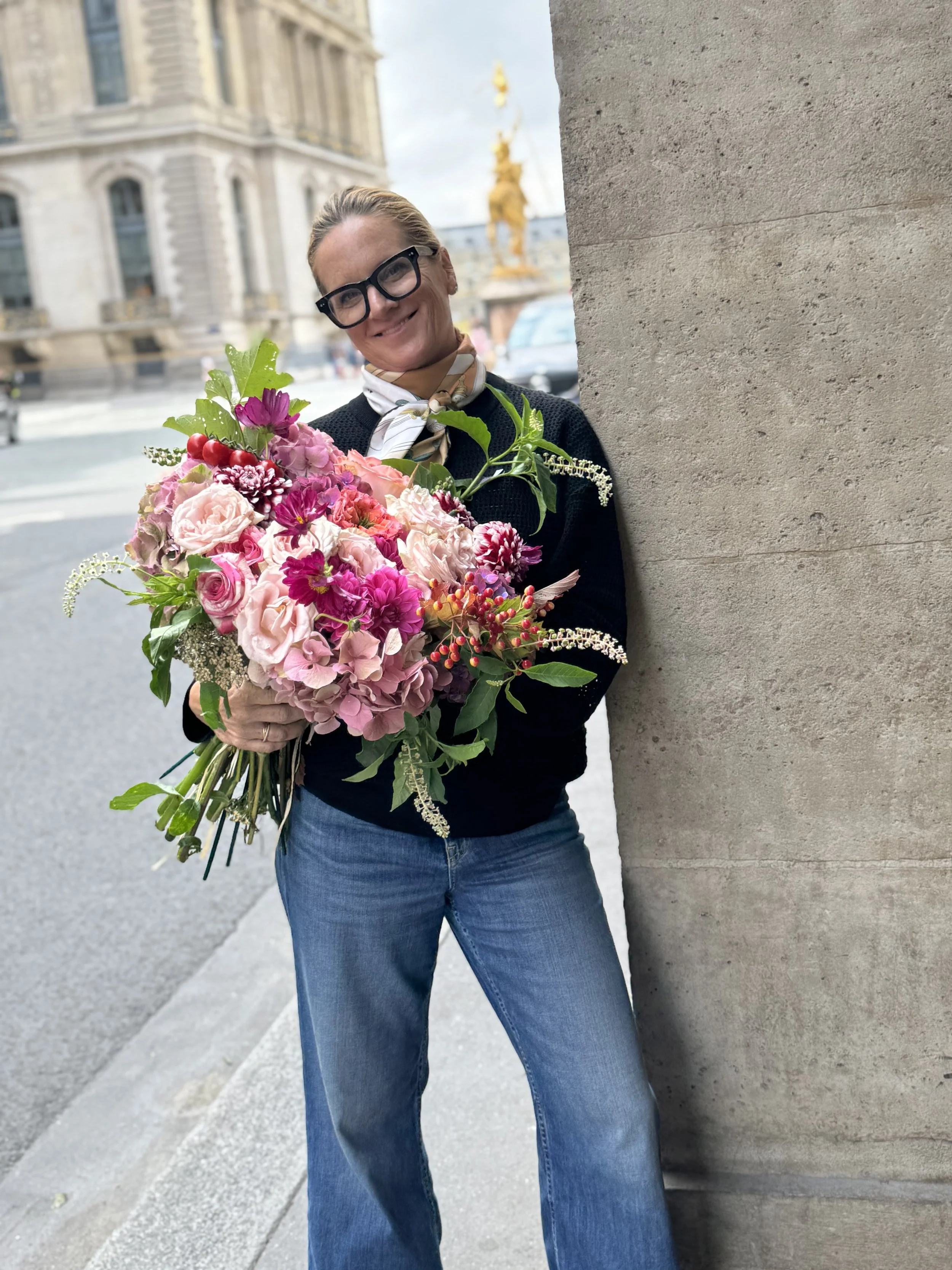 A woman with glasses and a scarf holding a large bouquet of pink, red, and purple flowers on a city sidewalk with historic buildings and a golden statue in the background.