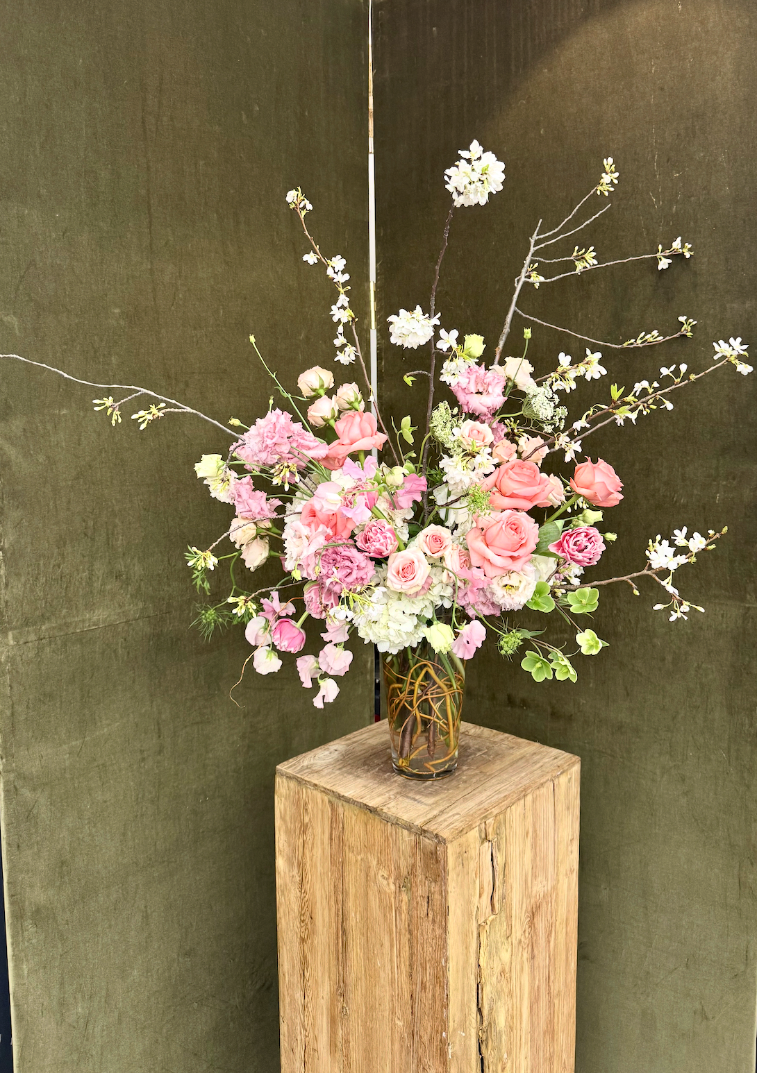 A tall floral arrangement with pink roses, white flowers, and branches in a glass vase, placed on a wooden pedestal against a dark background.