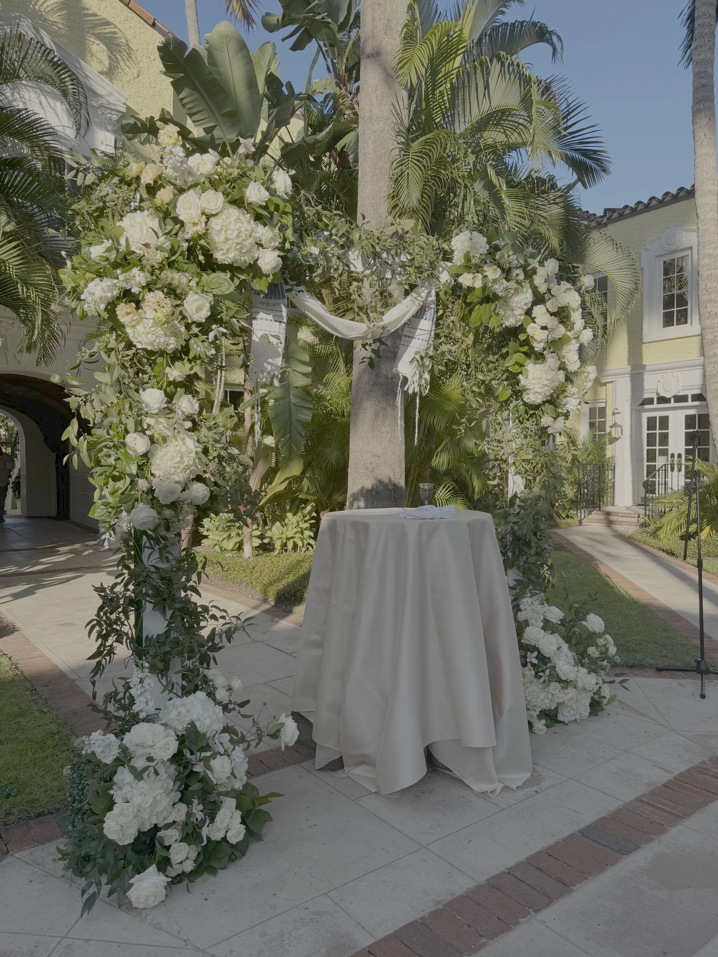 A decorated outdoor space with white flowers and greenery, including hydrangeas and roses, arranged around a small table with a white tablecloth, in front of a stone pillar and a house with yellow walls and palm trees.