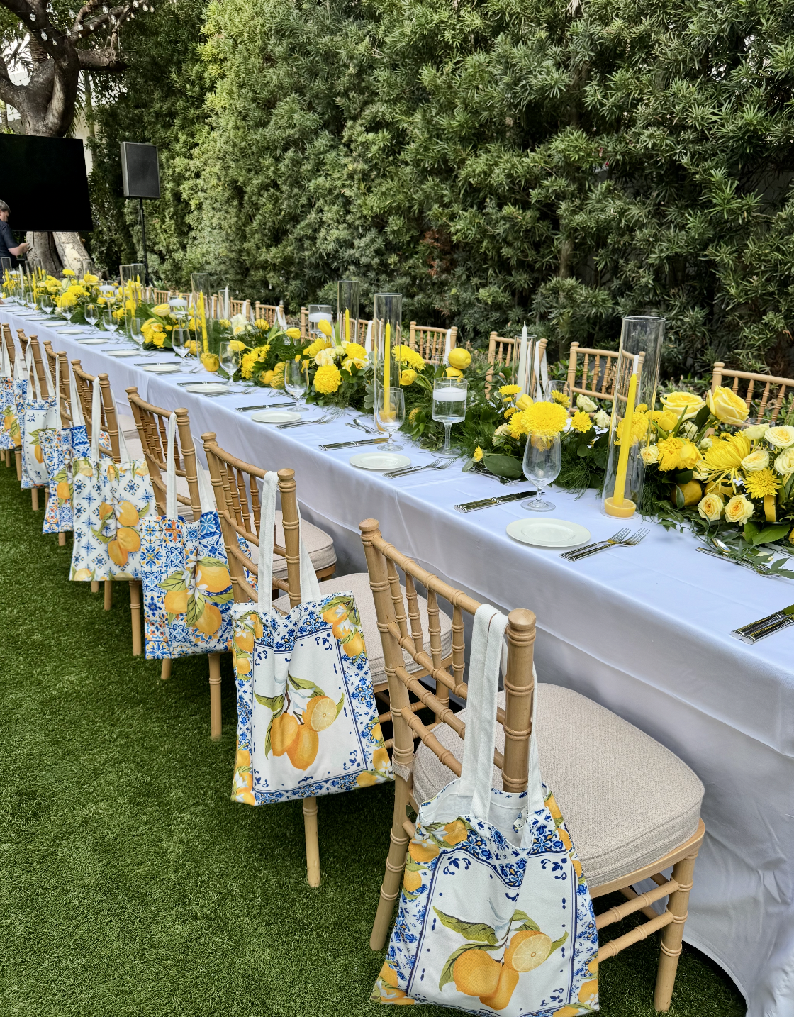 Long outdoor dining table decorated with yellow flowers, candles, and glassware, with chairs adorned with lemon-themed tote bags, set against green shrubbery.